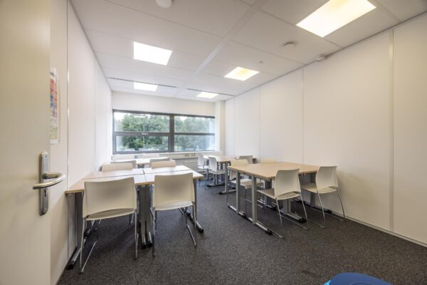 Kleine, moderne vergaderruimte of klaslokaal aan de Daltonlaan met witte muren, bureaus, stoelen en een groot raam met uitzicht op groen.
