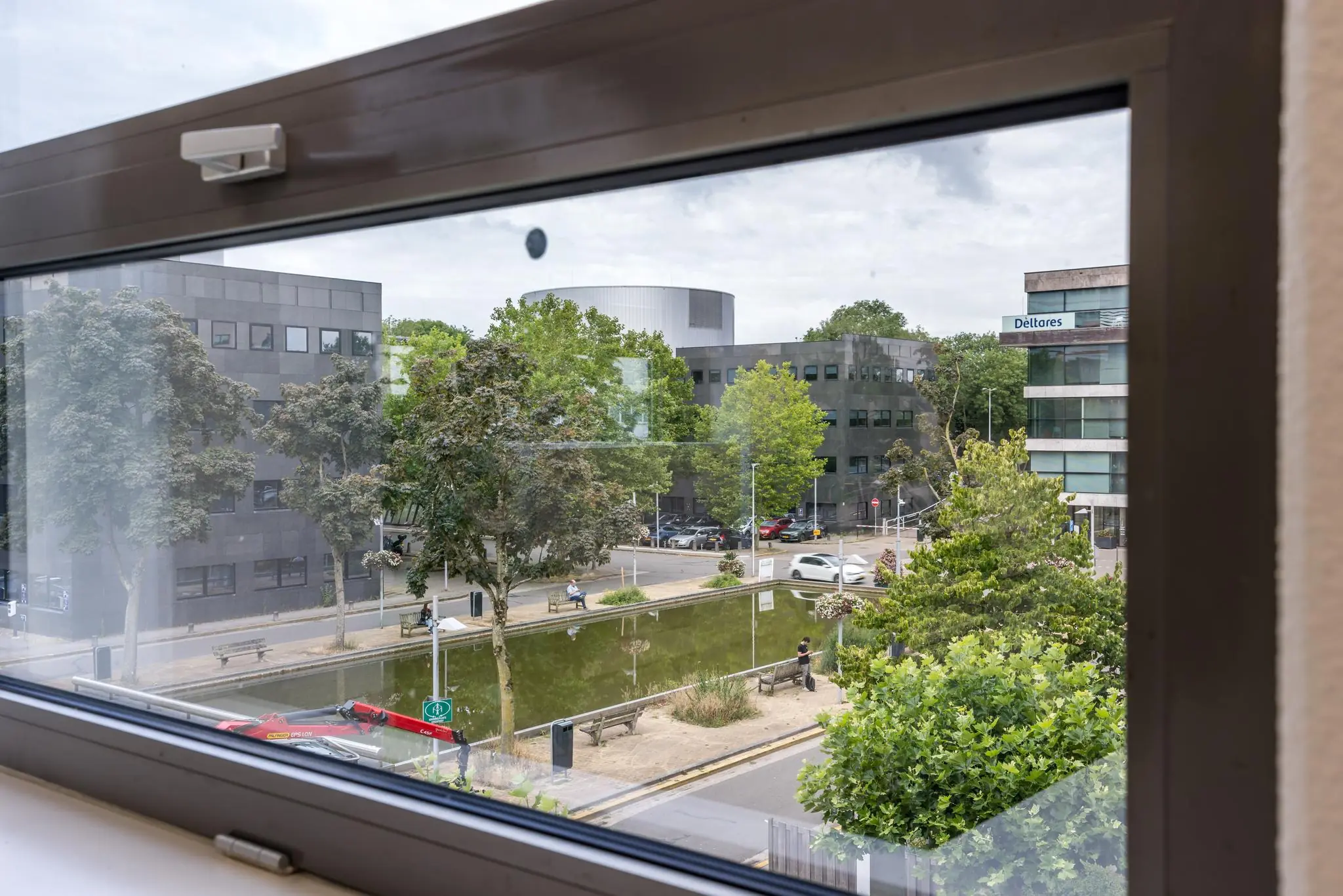 Uitzicht vanuit een raam op kantoorgebouwen aan de Daltonlaan met een vijver, groenstroken en het Deltares-gebouw op de achtergrond.