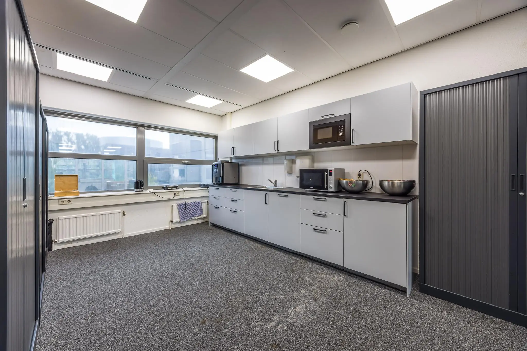 Moderne kantoorkeuken aan de Daltonlaan met grijze kasten, apparatuur en groot raam met daglicht.