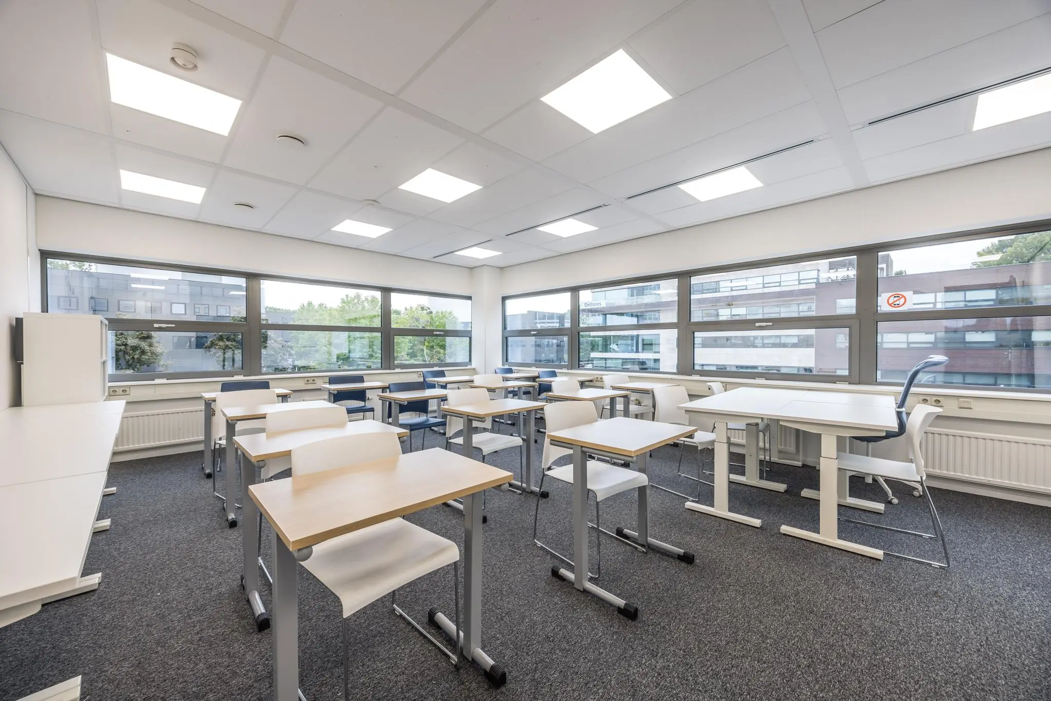 Modern ingerichte klaslokaal aan de Daltonlaan met witte bureaus, stoelen en grote ramen die uitzicht bieden op aangrenzende kantoorgebouwen.