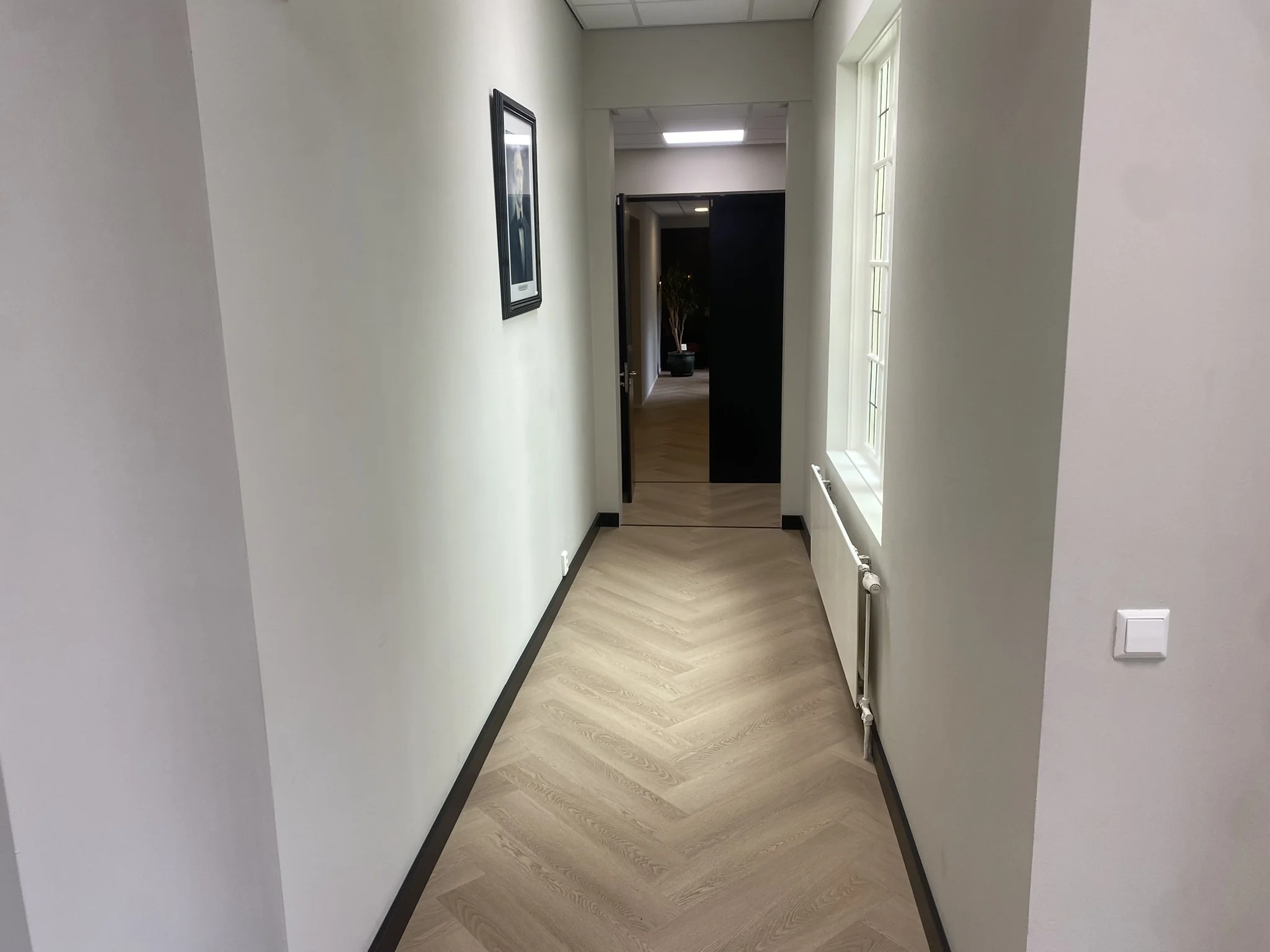 Verlicht gangpad in een kantoor aan het Lange Voorhout met houten visgraatvloer, witte muren en een raam aan de rechterkant.