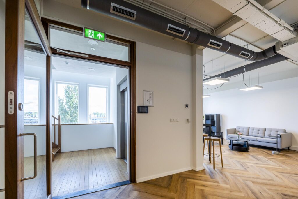 Modern ingerichte kantoorruimte aan de De Ruijterkade met houten vloer, zitgedeelte en zicht op een trap bij grote ramen.