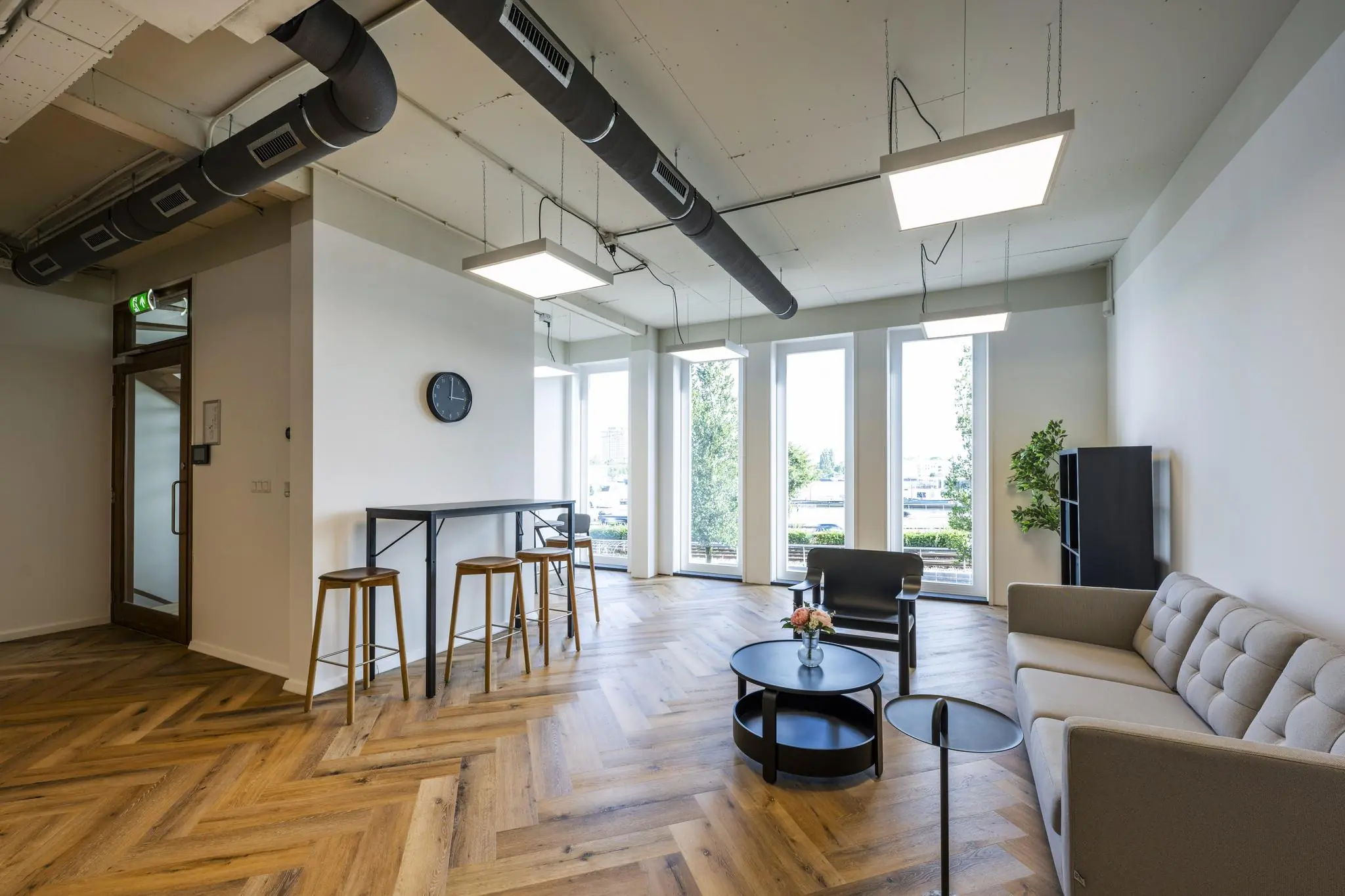 Moderne wachtruimte op De Ruijterkade met houten visgraatvloer, grote ramen, een zitgedeelte en hoge tafel met barkrukken.