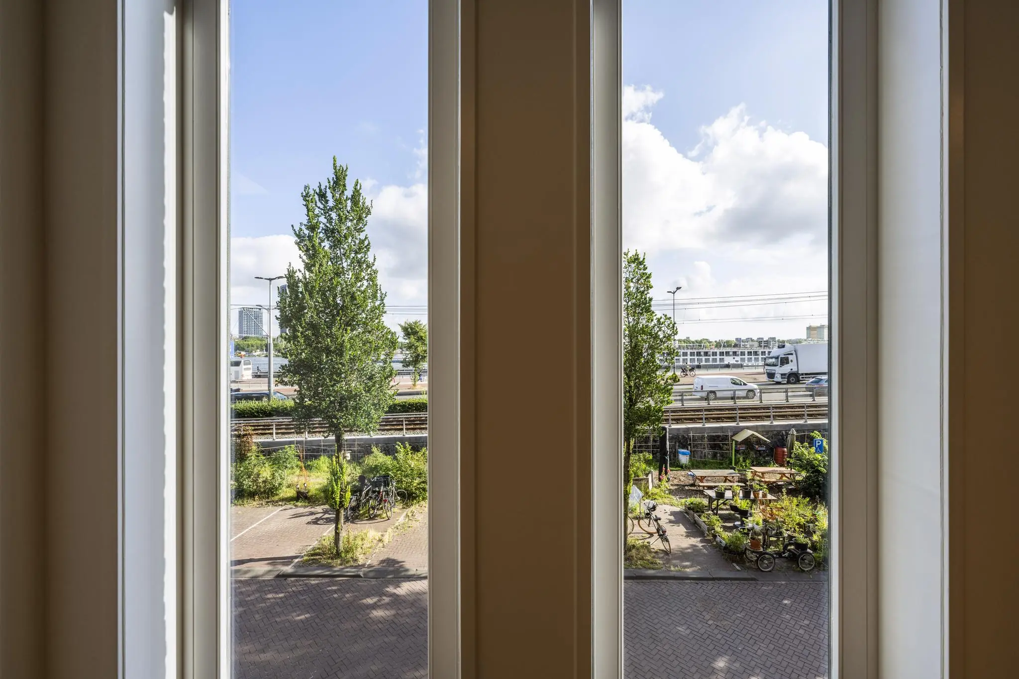 Uitzicht door drie hoge ramen op de De Ruijterkade met voorbijrijdend verkeer, groenstroken en een kleine stadstuin.