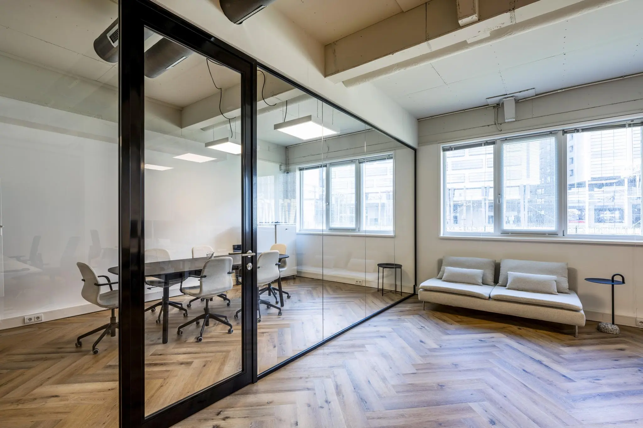 Modern kantoorinterieur aan de De Ruijterkade met glazen vergaderruimte, houten visgraatvloer en minimalistische zitplek.