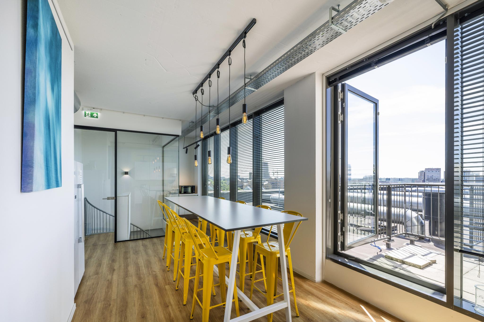 Moderne vergaderruimte met lange tafel, gele barkrukken en uitzicht op het Elizabeth Cady Stantonplein via een open glazen deur.