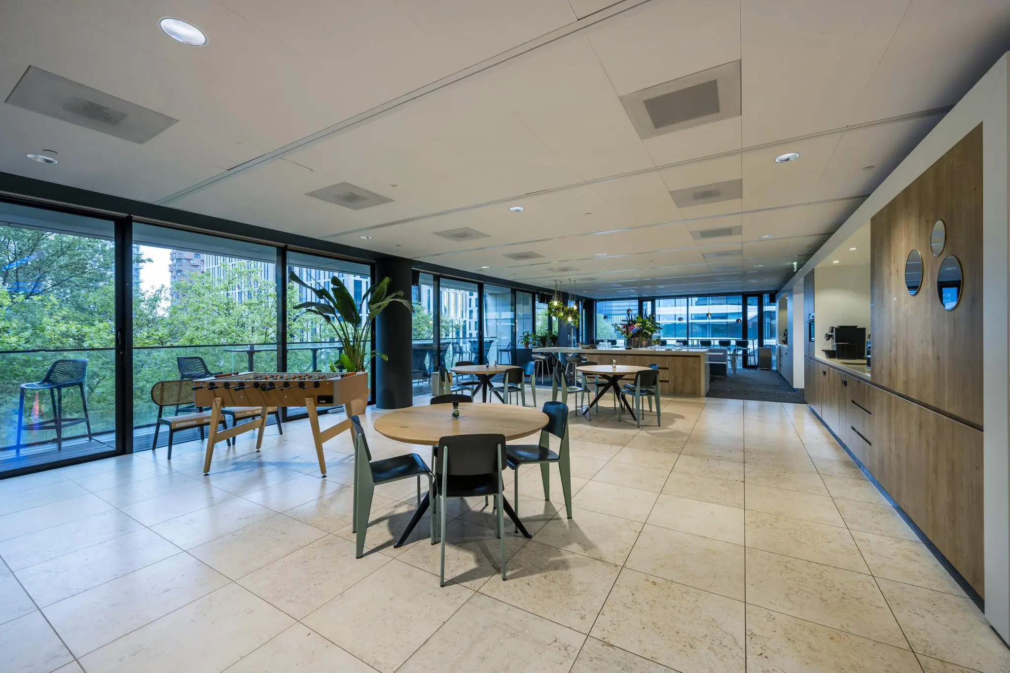 Moderne, open kantoorlounge aan de Gustav Mahlerlaan met zitplaatsen, een tafelvoetbaltafel en uitzicht op groen.