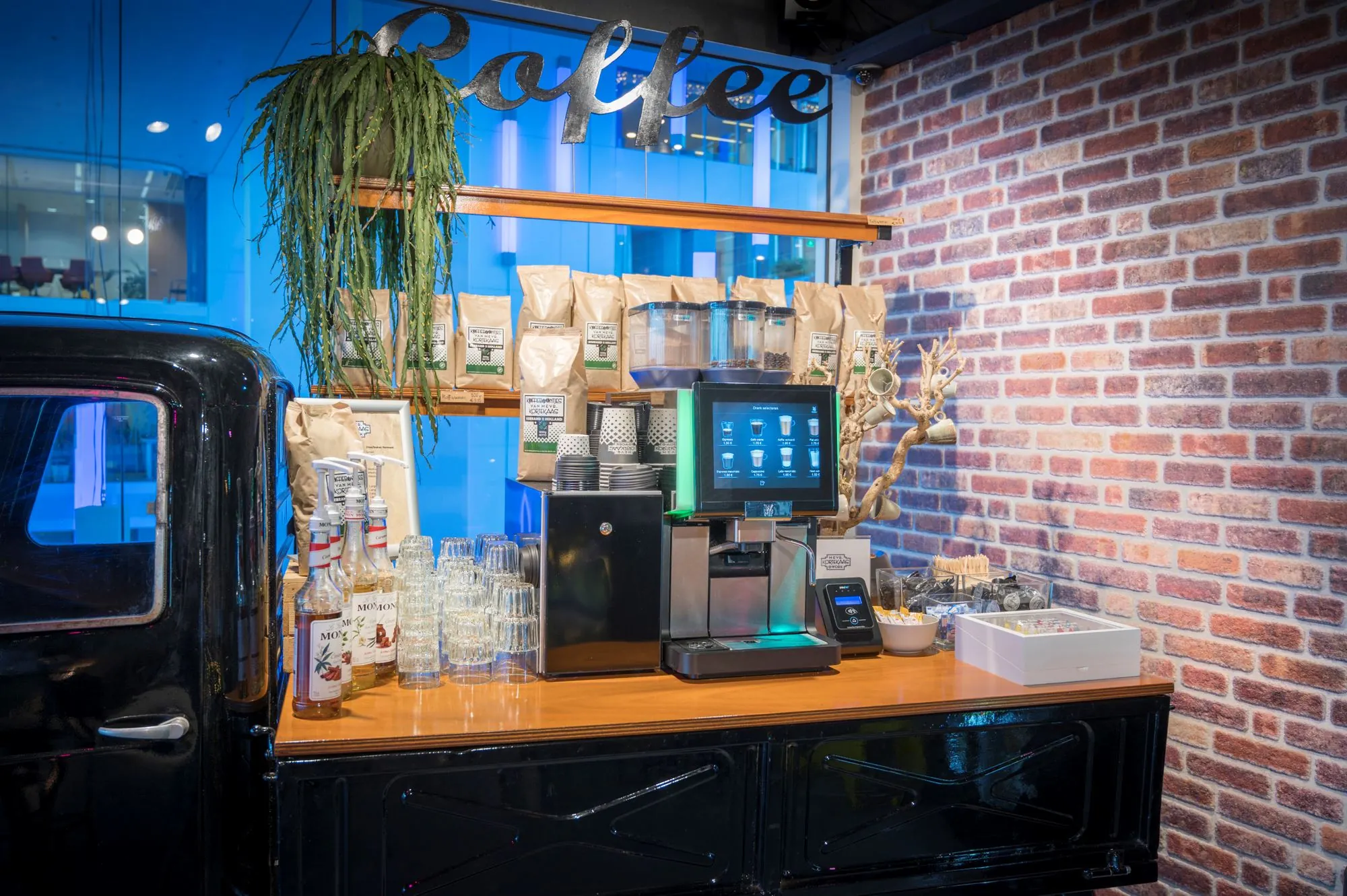 Koffiehoek met een zelfbedieningskoffiemachine, koffiebenodigdheden en decoratie tegen een bakstenen muur.