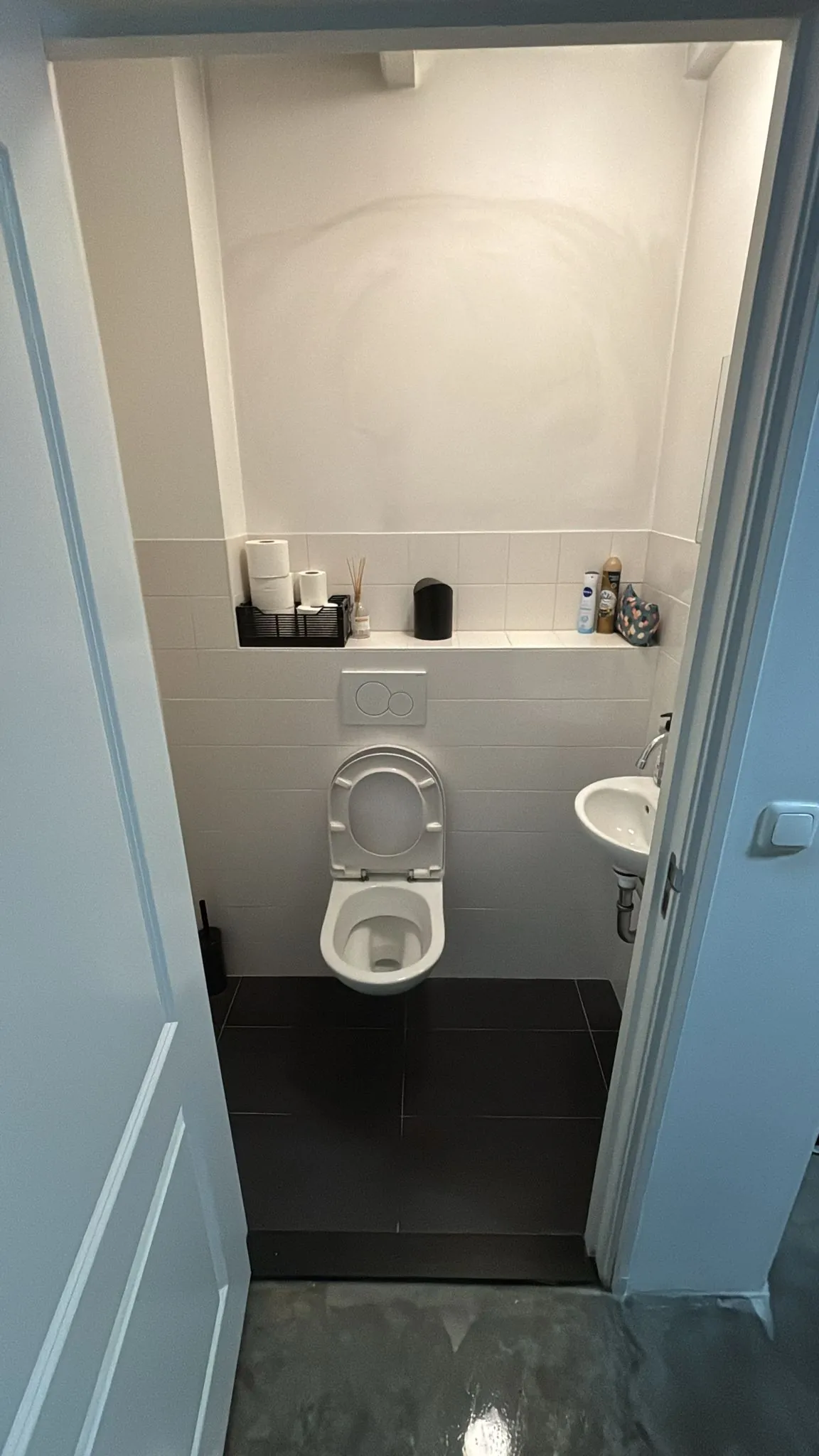 Moderne, smalle toiletruimte aan de Van Ostadestraat met wandcloset, zwart tegelvloer en een klein fonteintje.