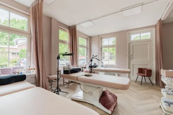 Lichtroze behandelkamer aan de Sloterweg met meerdere behandelbanken, grote ramen en zachte gordijnen.