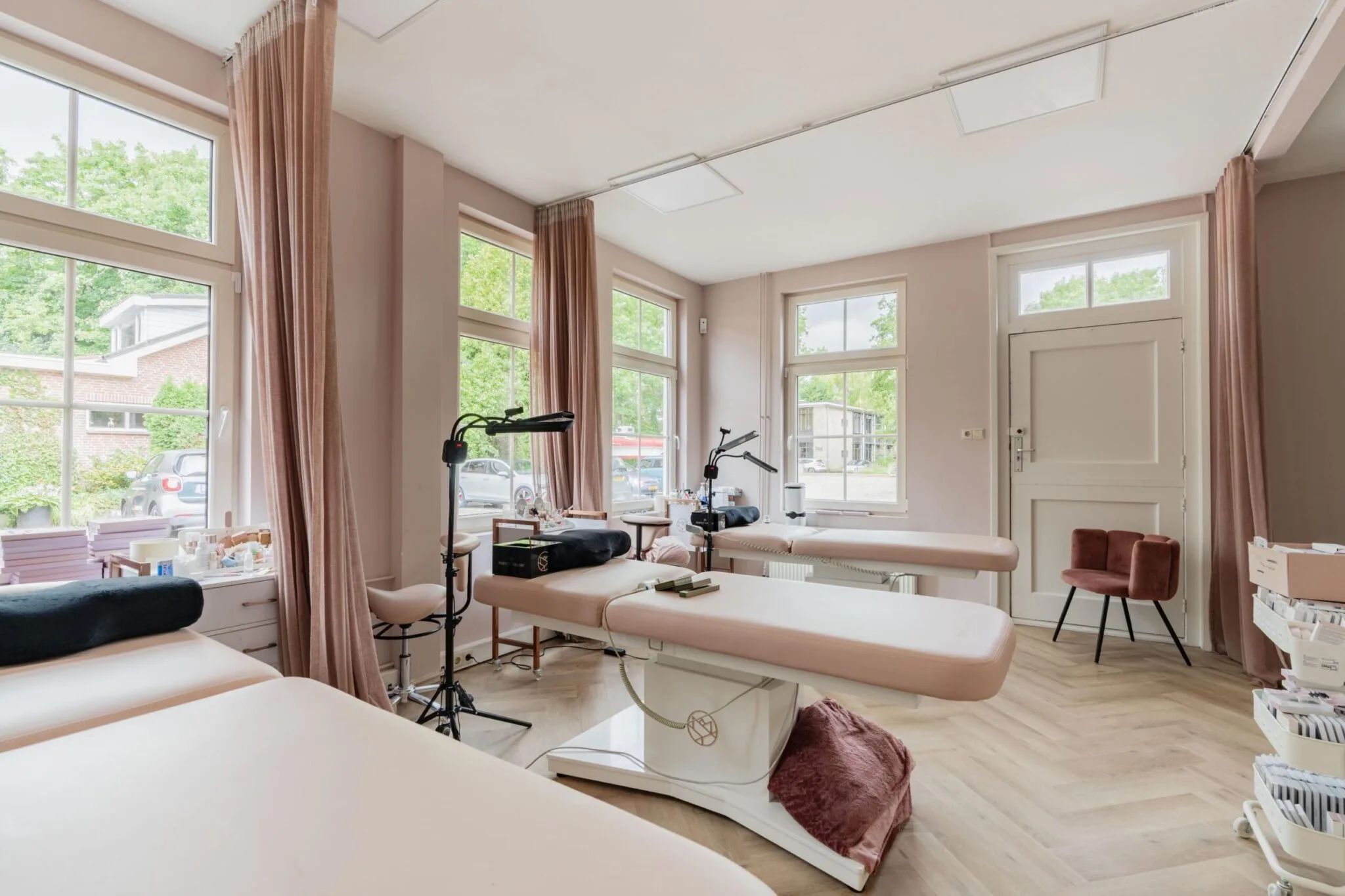 Lichtroze behandelkamer aan de Sloterweg met meerdere behandelbanken, grote ramen en zachte gordijnen.