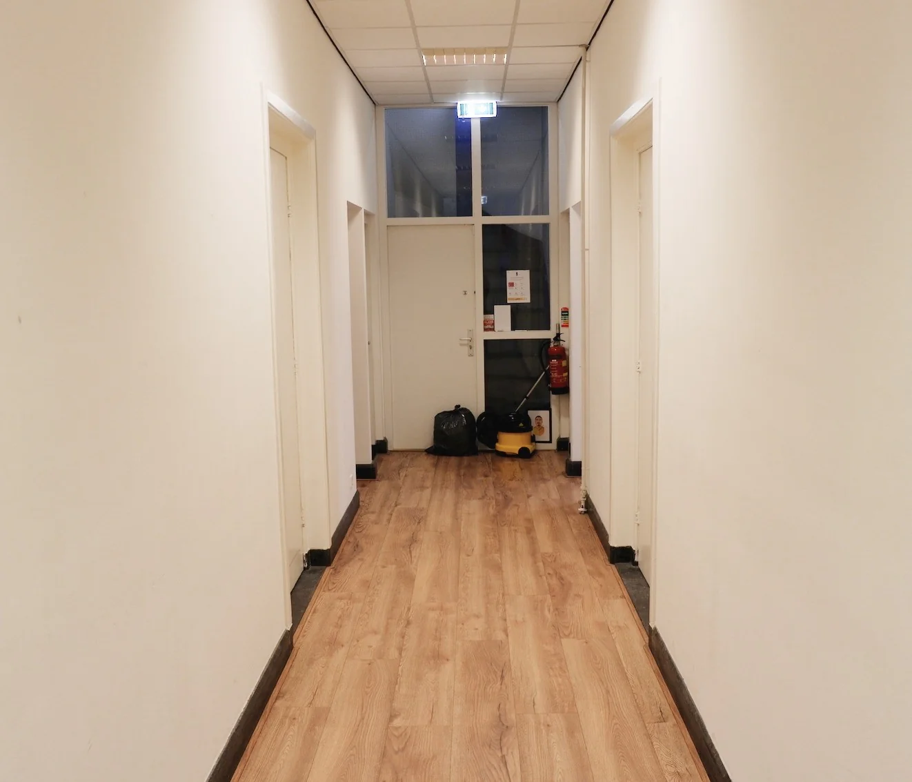 Verlichte gang met houten vloer in een gebouw aan de Raadhuisstraat, met schoonmaakspullen en een brandblusser aan het einde.