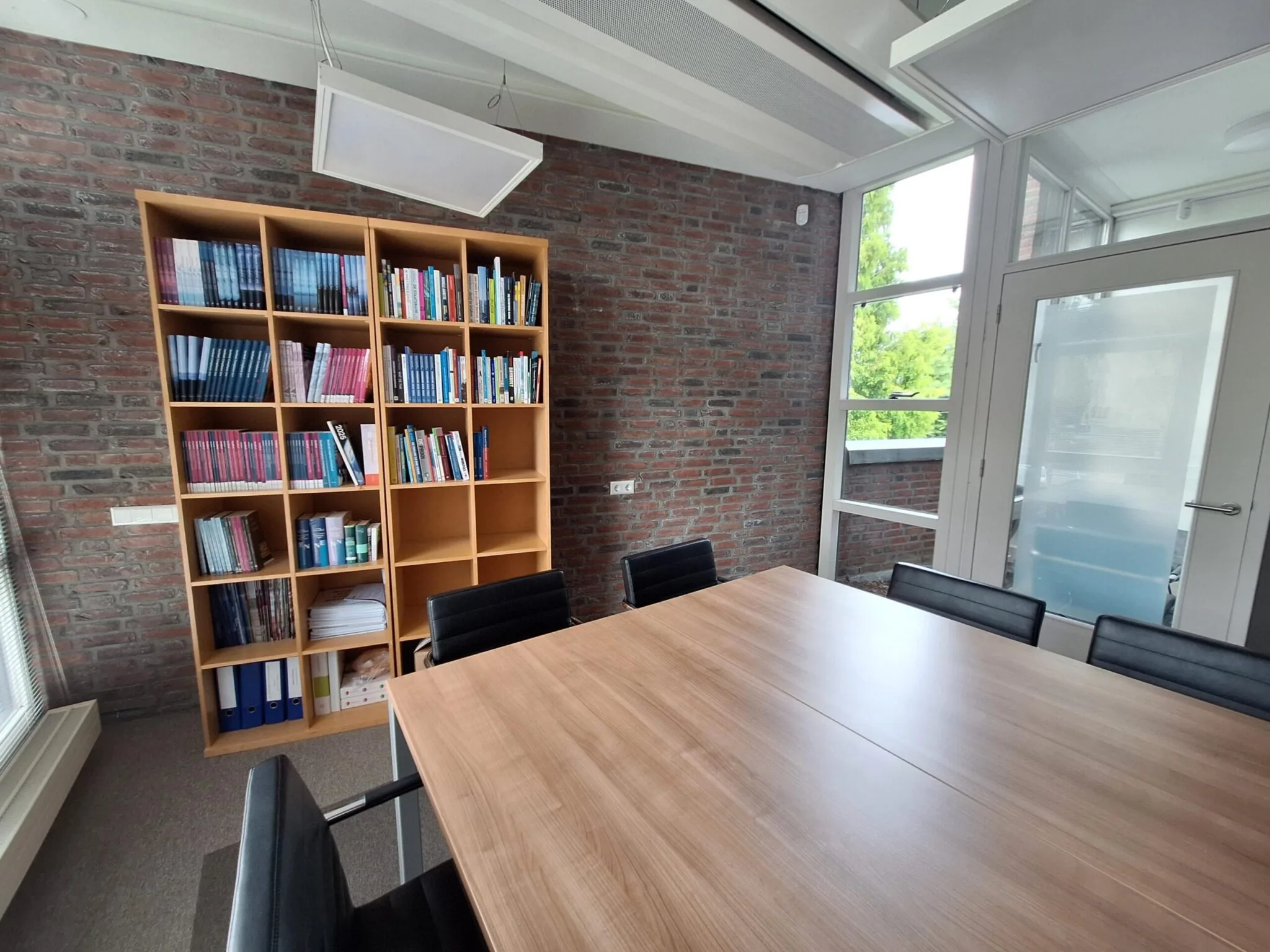 Vergaderruimte aan de Jan Leentvaarlaan met een houten tafel, zwarte stoelen en een gevulde boekenkast tegen een bakstenen muur.
