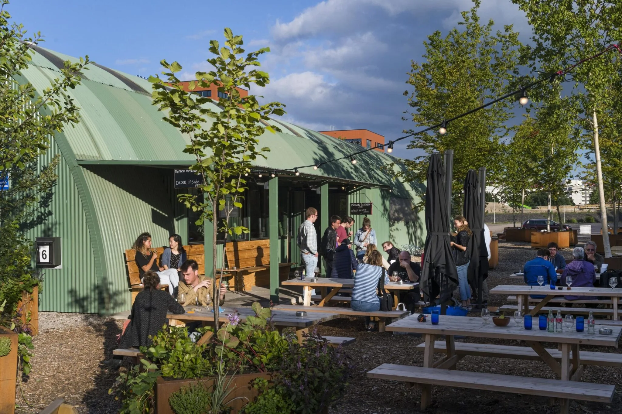 Terras met houten picknicktafels en mensen bij een groen gebouw aan de Kuiperbergweg op een zonnige dag.