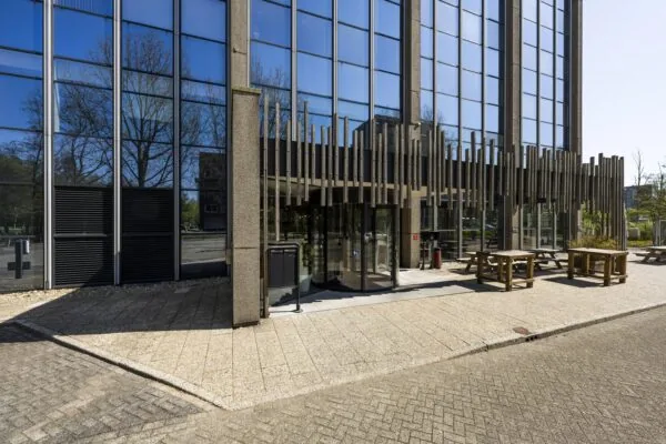 Ingang van een modern kantoorgebouw aan de Laan van Kronenburg met glazen gevel en zitbanken buiten.