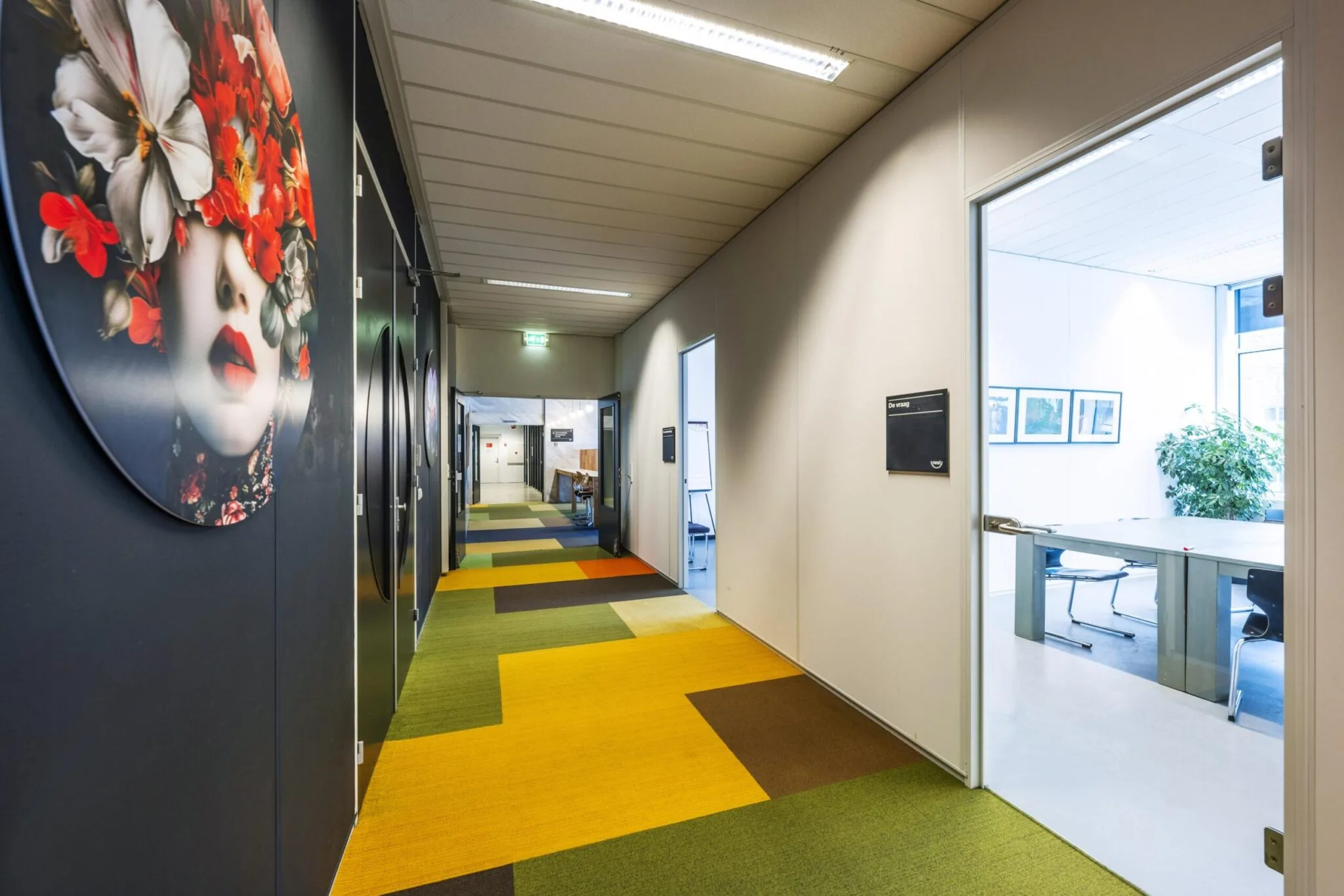 Kleurvolle gang in kantoorpand aan de Laan van Kronenburg met kunst aan de muur en zicht op vergaderruimte.