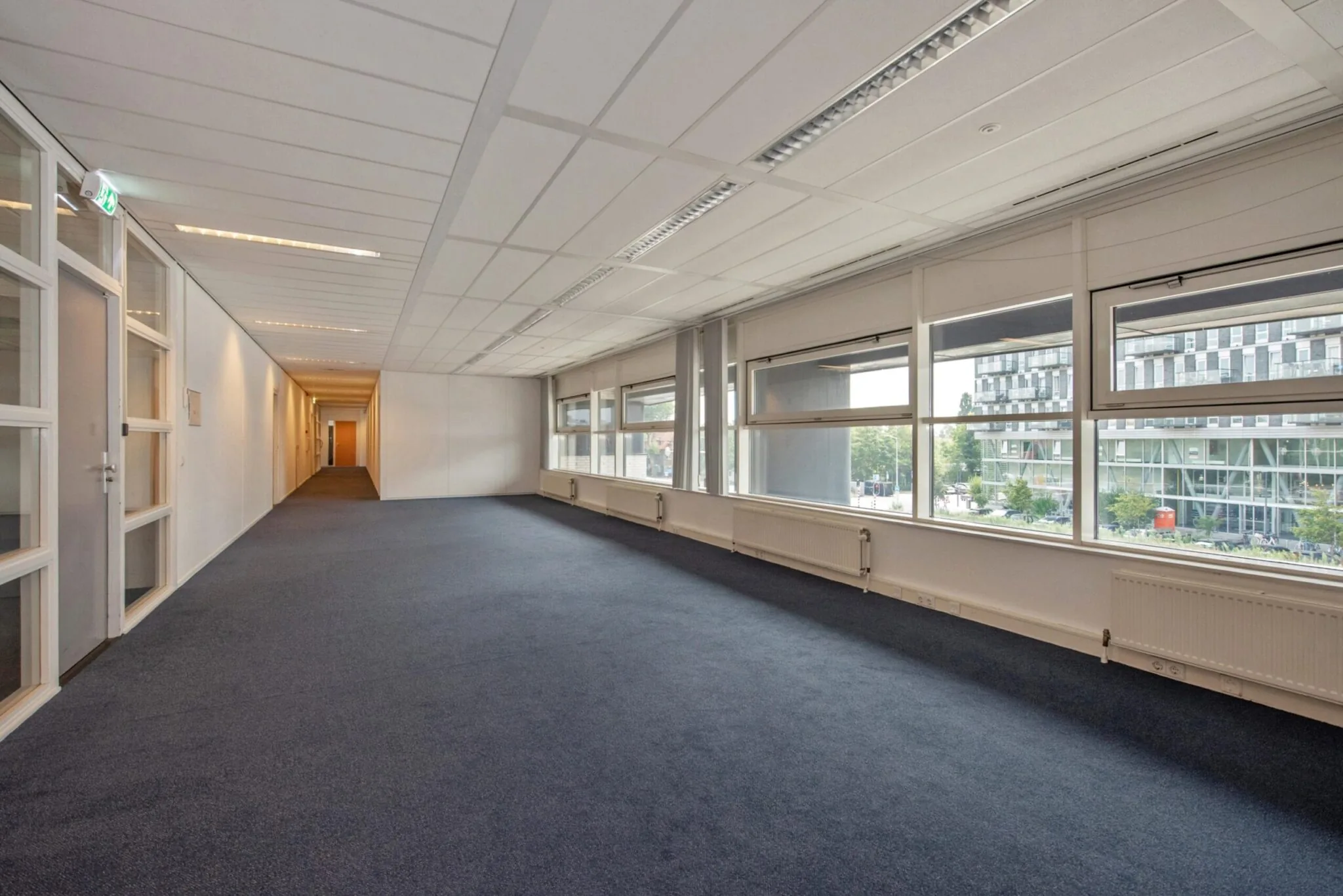Lichte, lege kantoorruimte aan de Binckhorstlaan met grote ramen en donkerblauw tapijt.