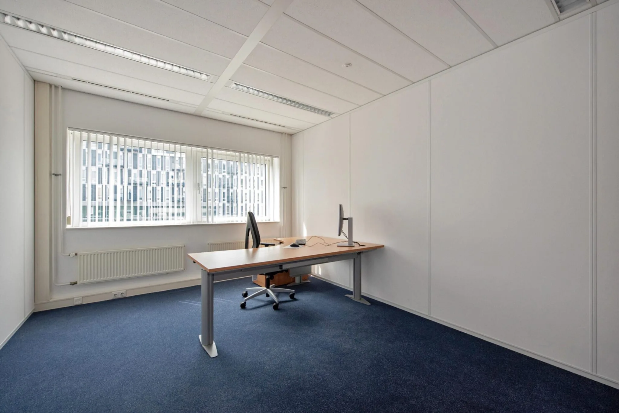 Kantoorruimte aan de Binckhorstlaan met een enkel bureau, bureaustoel en beeldscherm, gelegen in een lichte kamer met grote ramen en verticale lamellen.