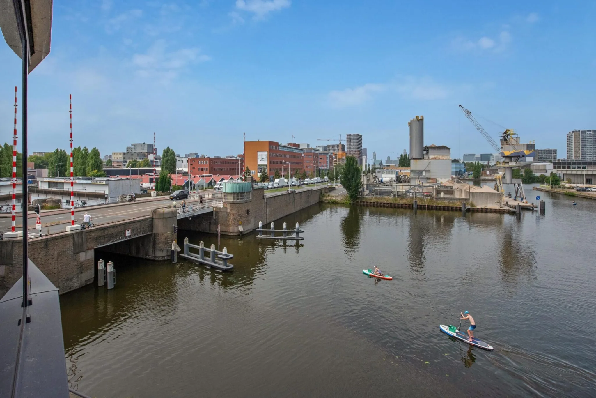 Uitzicht op de Binckhorstlaan in Den Haag met een brug over het water, industriegebouwen en twee suppers op het kanaal.