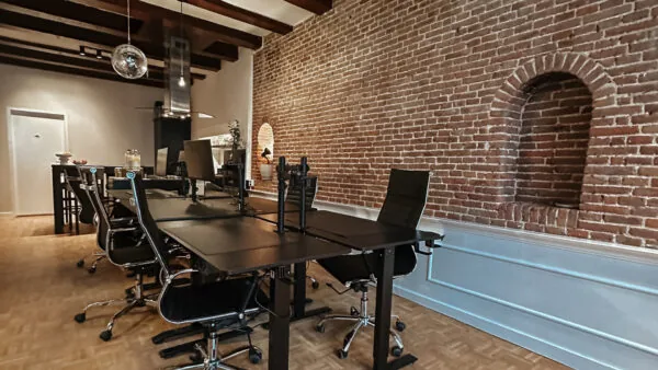 Moderne kantoorruimte aan de Herengracht met zwarte bureaus en stoelen, een bakstenen muur en zichtbare houten plafondbalken.