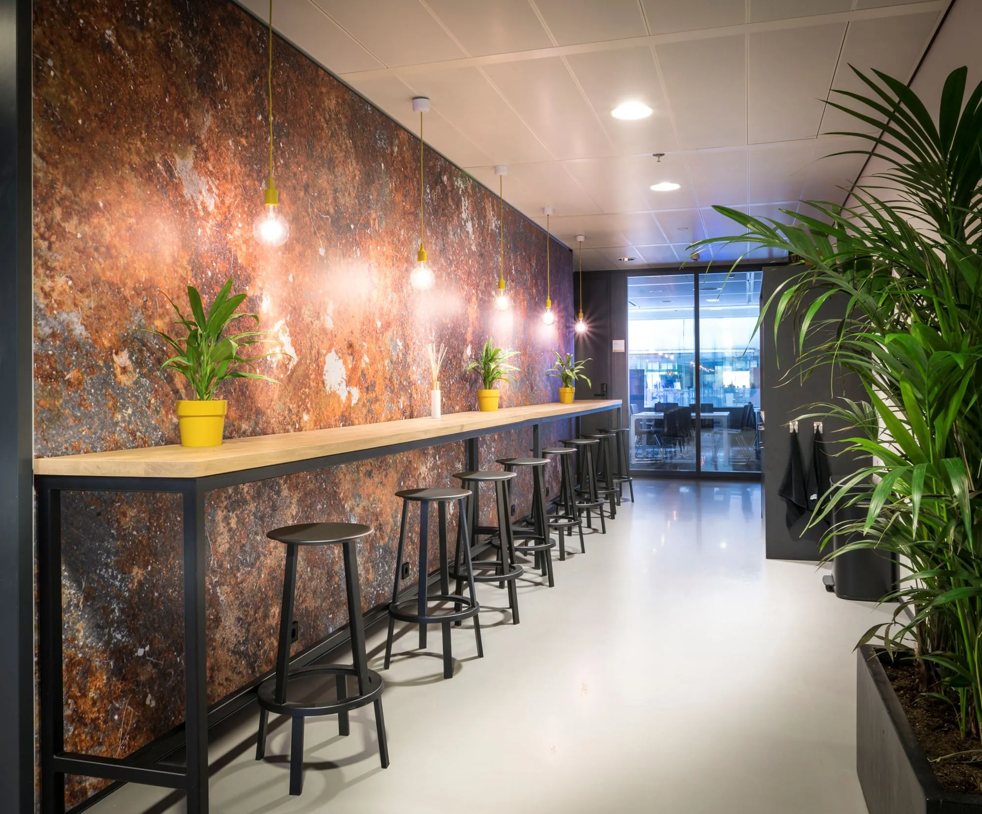 Moderne kantoorruimte met een hoge houten bar, zwarte barkrukken, hanglampen en groene planten in gele potten.