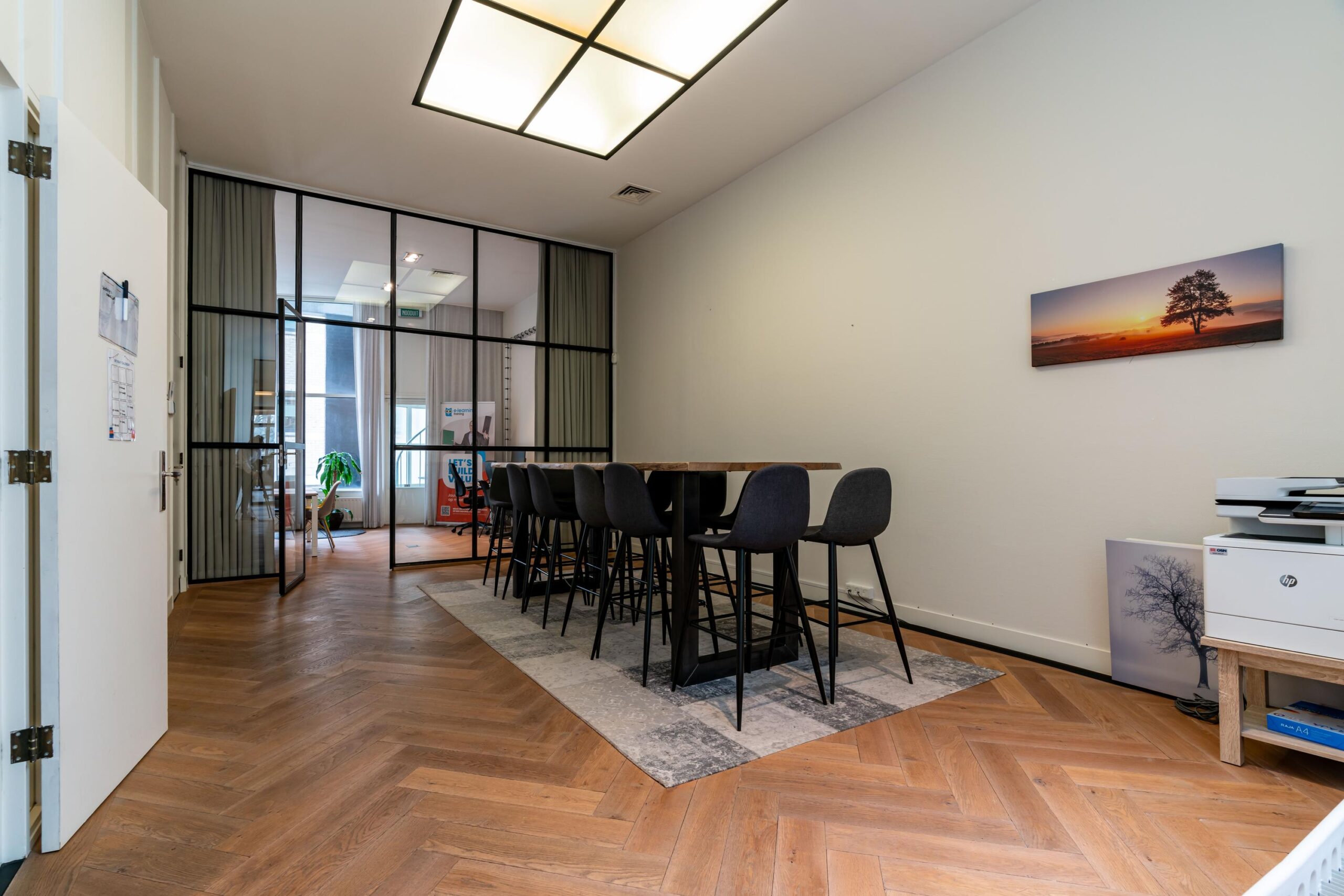 Vergaderruimte aan de Lange Vijverberg met hoge tafel, barkrukken, houten visgraatvloer en glazen wand naar aangrenzend kantoor.
