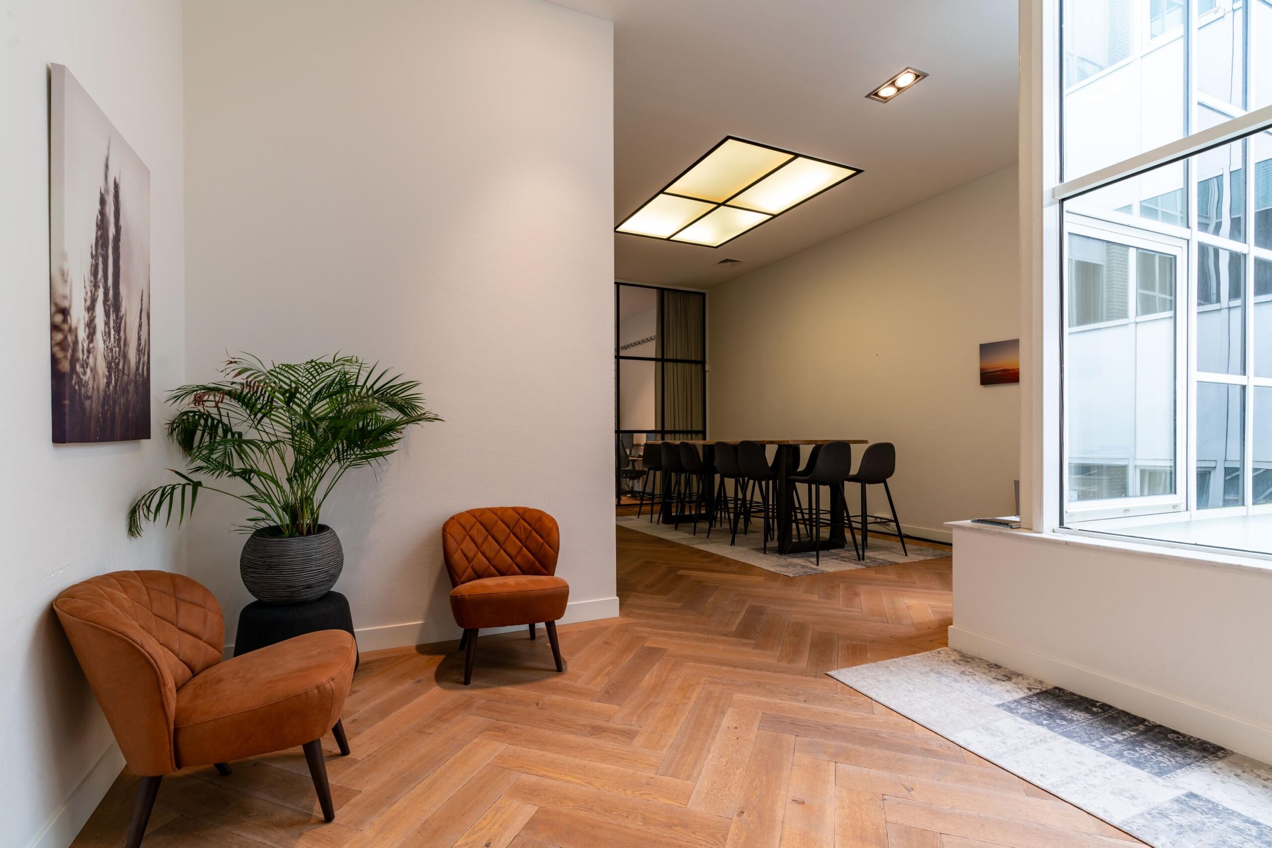 Moderne wachtruimte met twee bruine stoelen, een grote plant en uitzicht op een vergaderruimte met hoge stoelen aan de Lange Vijverberg.
