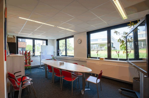 Vergaderruimte aan de Veteranenlaan met ovale tafel, rode stoelen, whiteboard en uitzicht op moderne kantoorgebouwen.