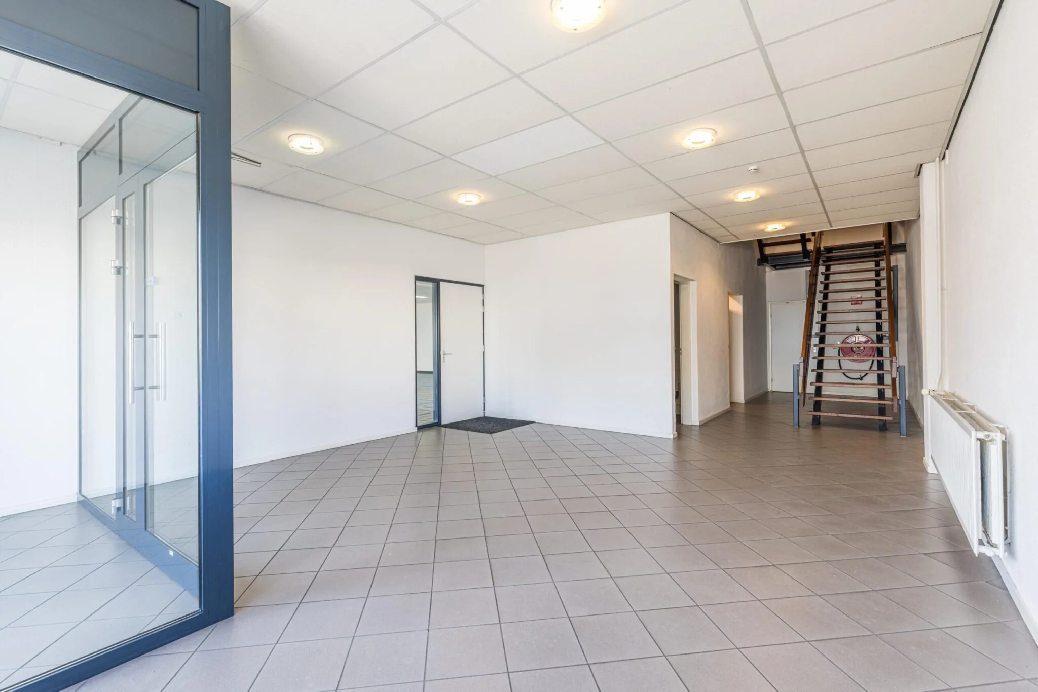 Lichte entreehal met tegelvloer, glazen scheidingswand en houten trap in een bedrijfsgebouw aan de Zonnebaan.