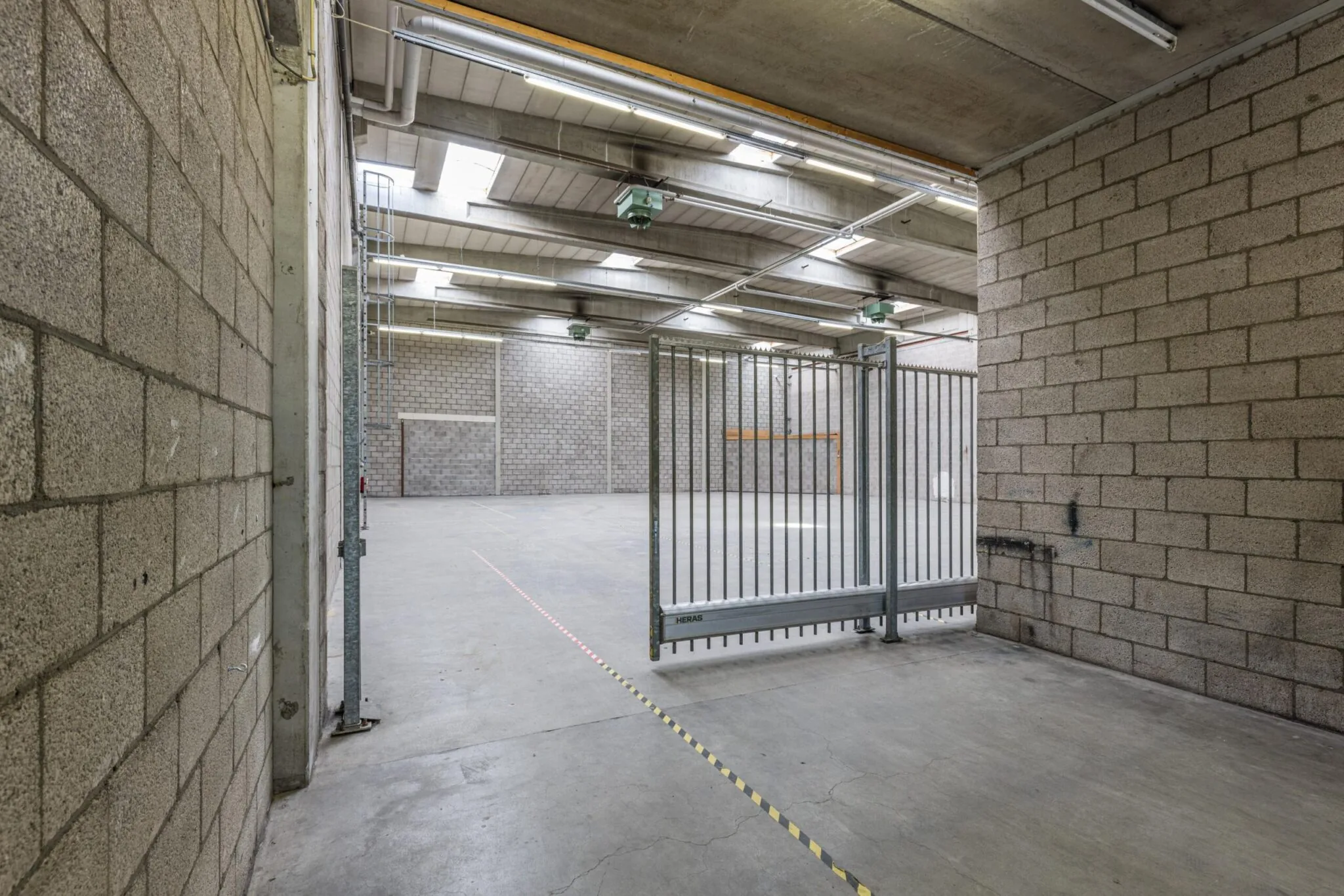 Ruime, lege bedrijfshal aan de Zonnebaan met betonnen vloer, hekwerk en zicht op hoge plafonds met verlichting.