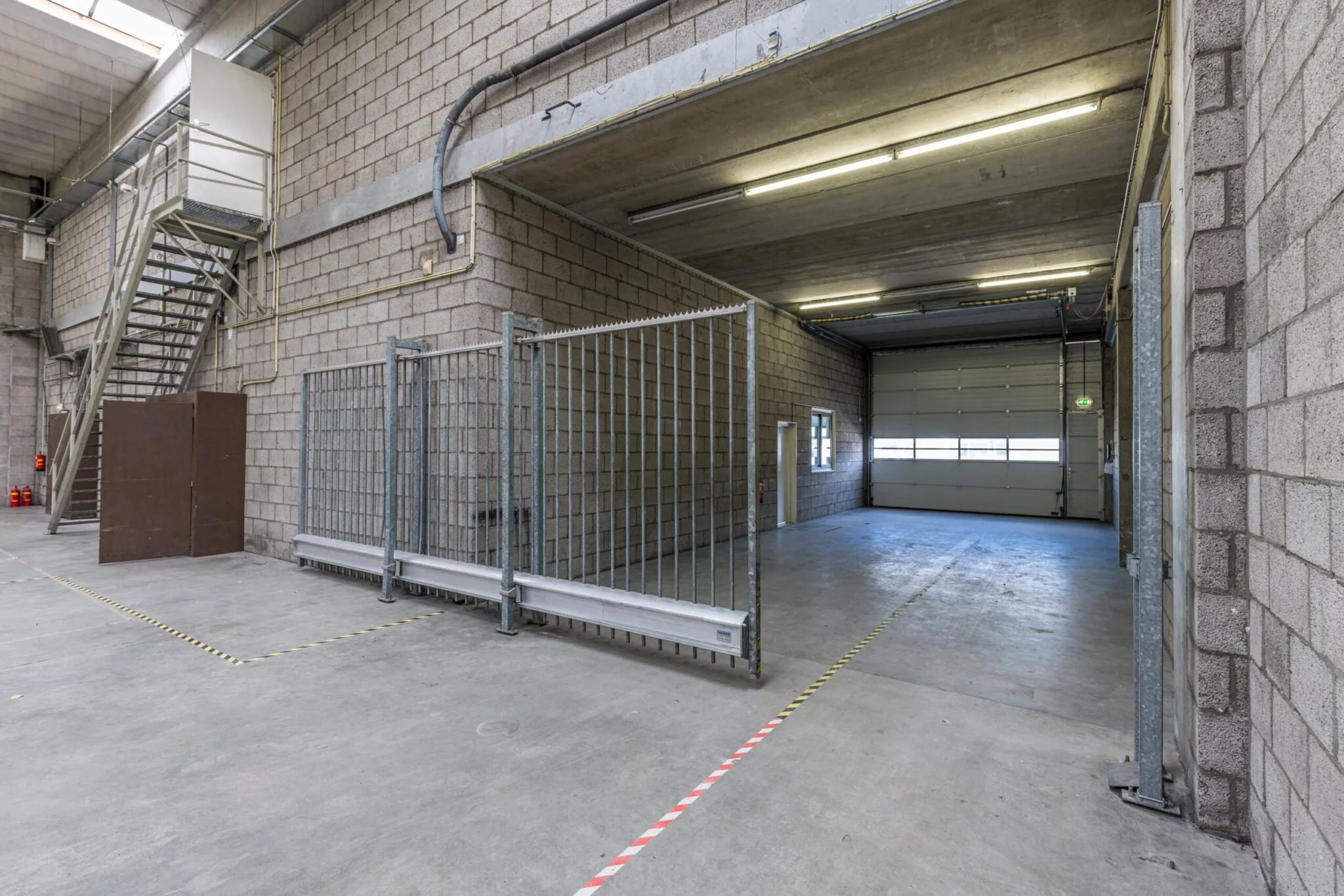 Ruime, lege bedrijfsruimte aan de Zonnebaan met betonnen vloeren, rolpoort en metalen trap.