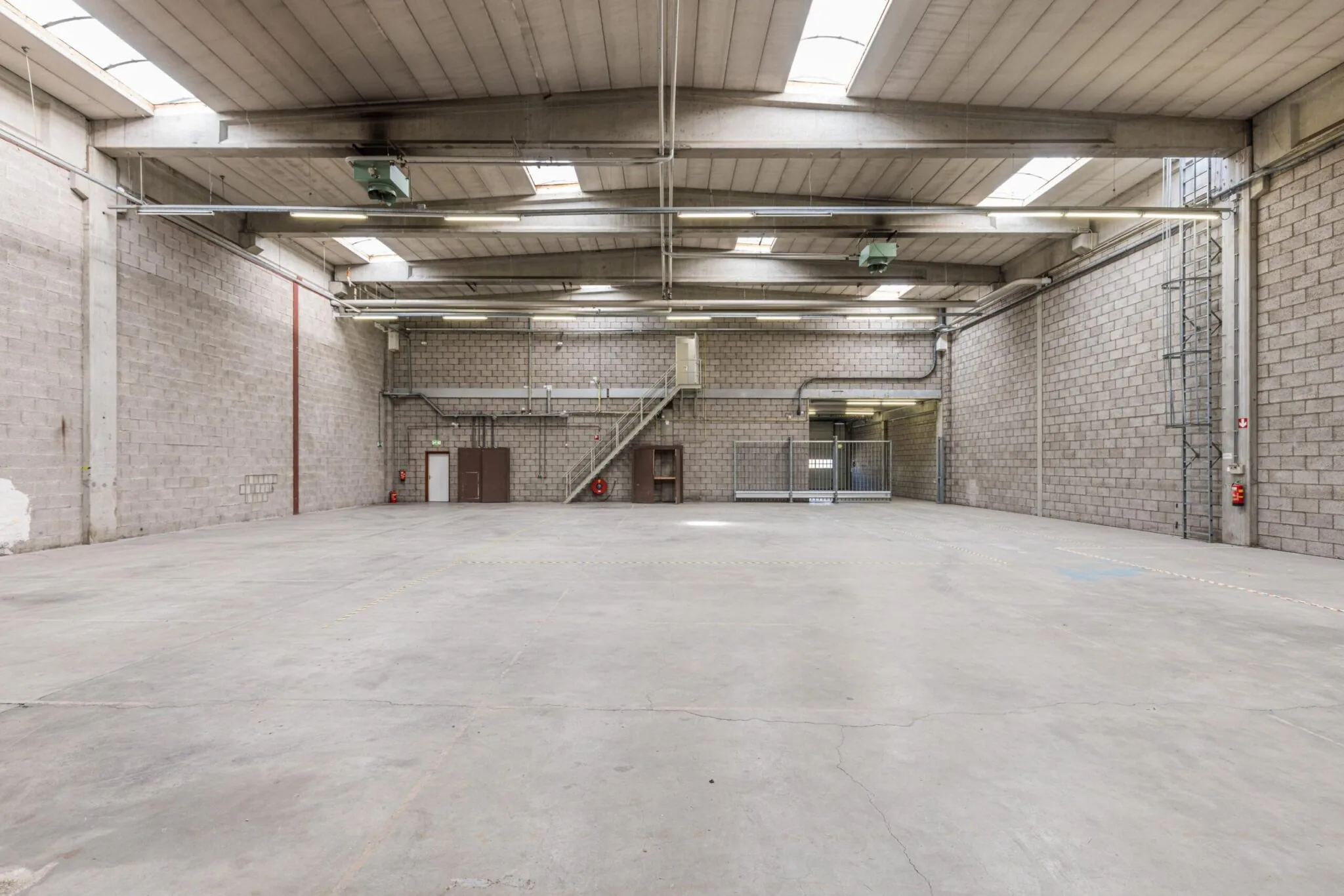 Ruime, lege bedrijfsruimte met betonnen vloer en zicht op metalen trap en grote toegangspoorten aan de Zonnebaan.