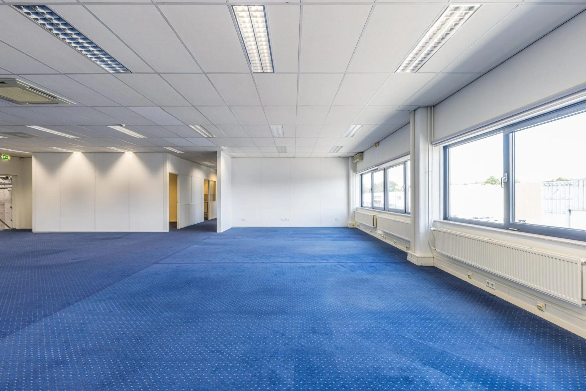 Lichte, leegstaande kantoorruimte aan de Zonnebaan met blauw tapijt en grote ramen.