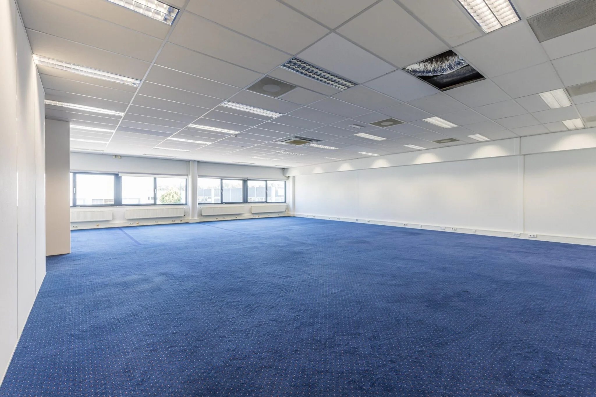 Lege kantoorruimte met blauw tapijt, systeemplafond en grote ramen aan de Zonnebaan.