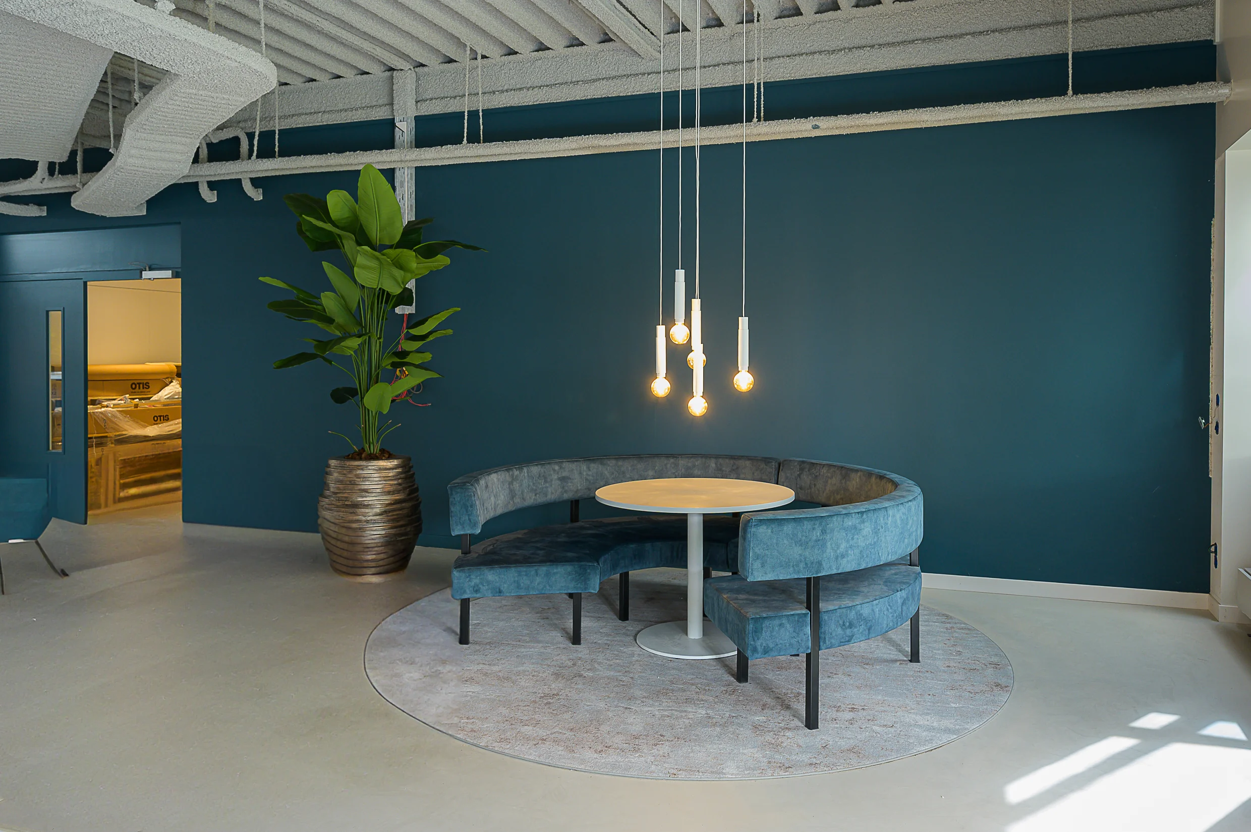 Modern lounge area with a round blue velvet bench, circular rug, hanging pendant lights, and a large potted plant against a dark teal wall.