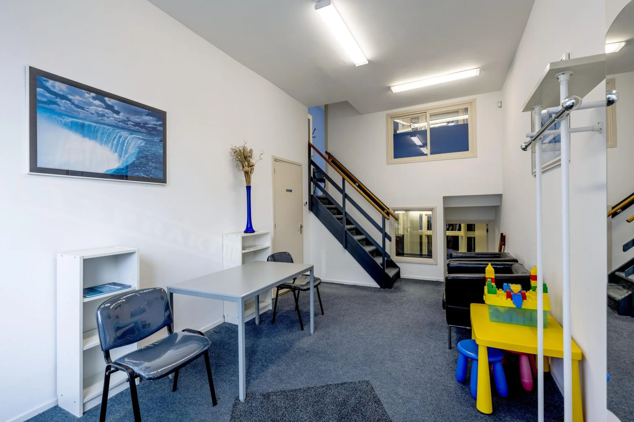 Wachtruimte met een trap, zitplaatsen, kinderspeelplek en een groot waterval-schilderij aan de muur.