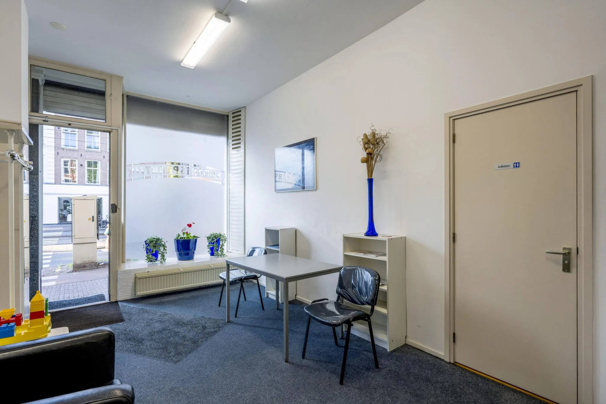 Wachtruimte met tafel en stoelen in een praktijk aan de Overtoom, met zicht op de straat door het raam.