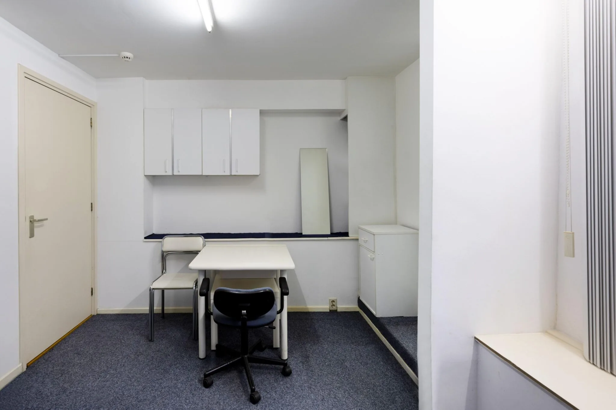 Kleine, eenvoudig ingerichte werk- of vergaderruimte met twee stoelen, een tafel, een spiegel en witte kasten aan de muur.
