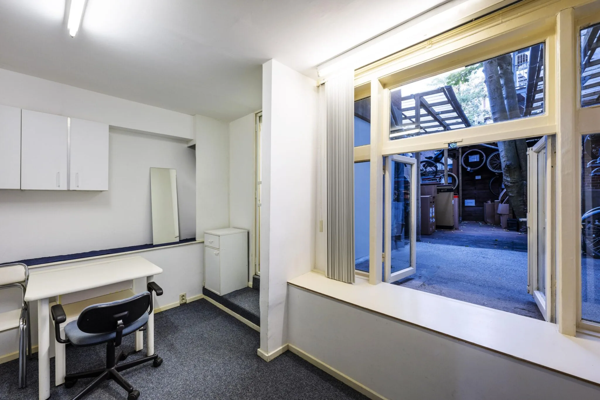 Eenvoudig ingericht kantoor of studeerkamer met een bureau en stoel, grenzend aan een binnentuin met fietsenrek aan de Overtoom.