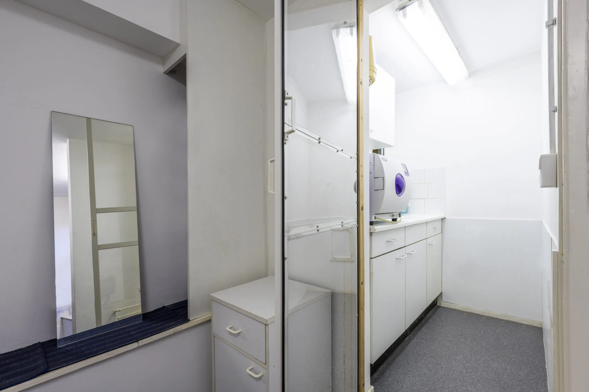 Kleine witte behandel- of sterilisatieruimte met een kast, spiegel en medisch apparaat op het aanrecht.