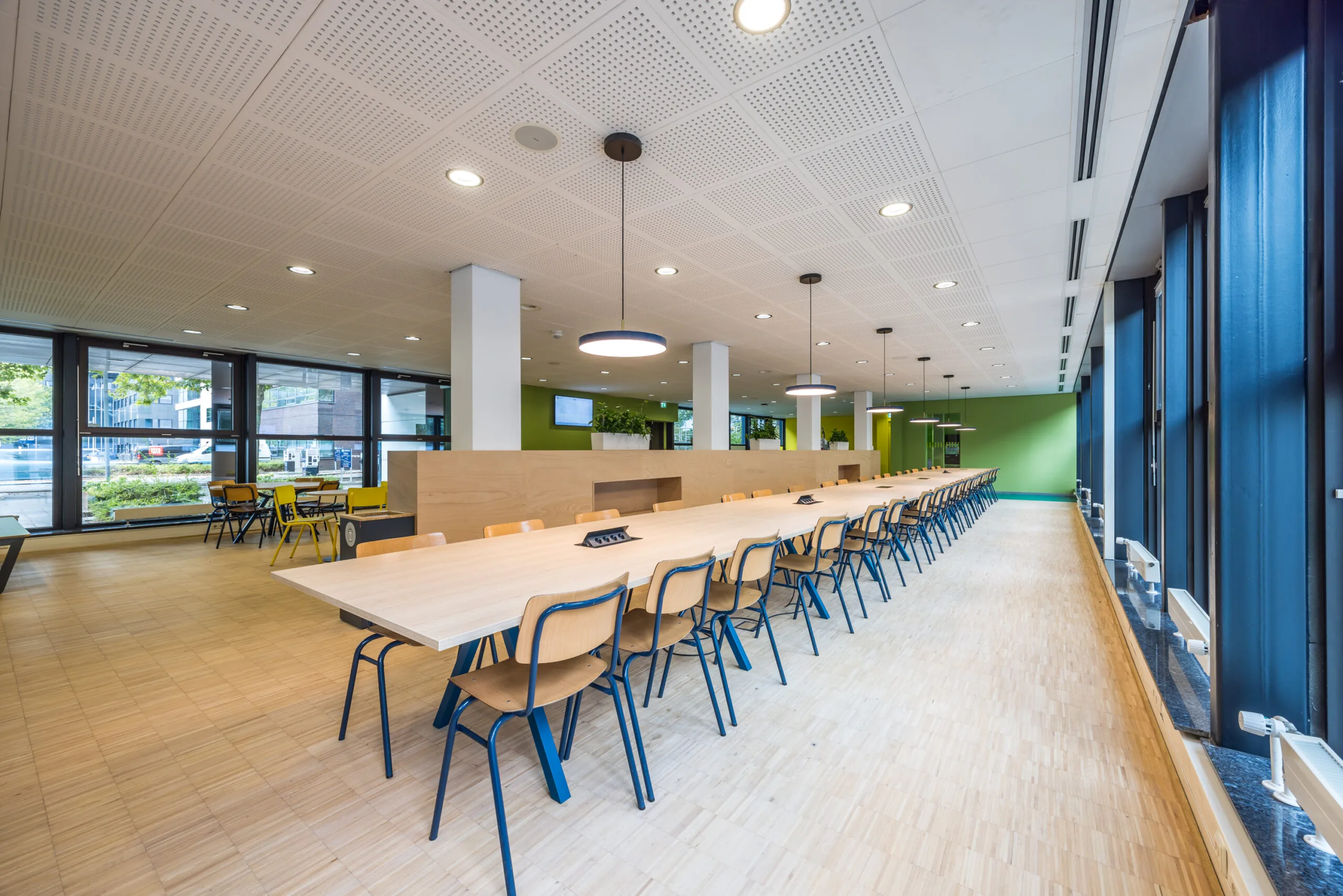 Moderne en ruim opgezette kantoorkantine aan de Daltonlaan met lange houten tafels, blauwe stoelen en grote ramen.