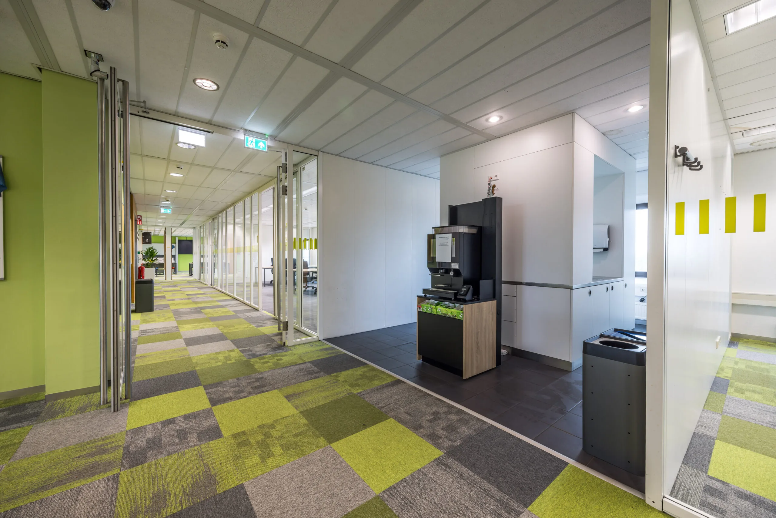 Moderne kantoorruimte aan de Daltonlaan met kleurrijke tapijttegels, glazen wanden en een koffieautomaat in de gang.