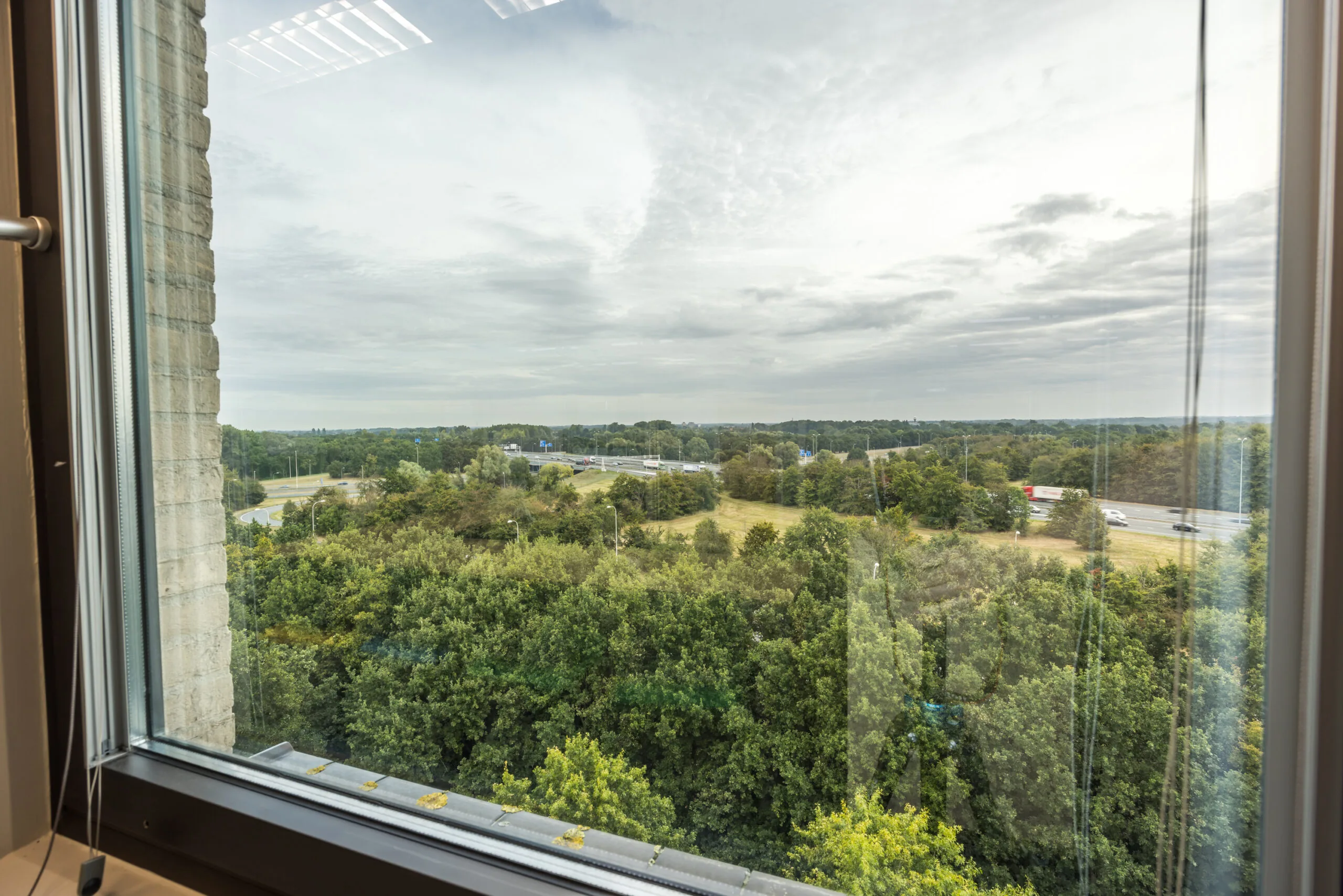 Uitzicht vanuit een raam op Daltonlaan met een snelweg, groen landschap en een bewolkte lucht.