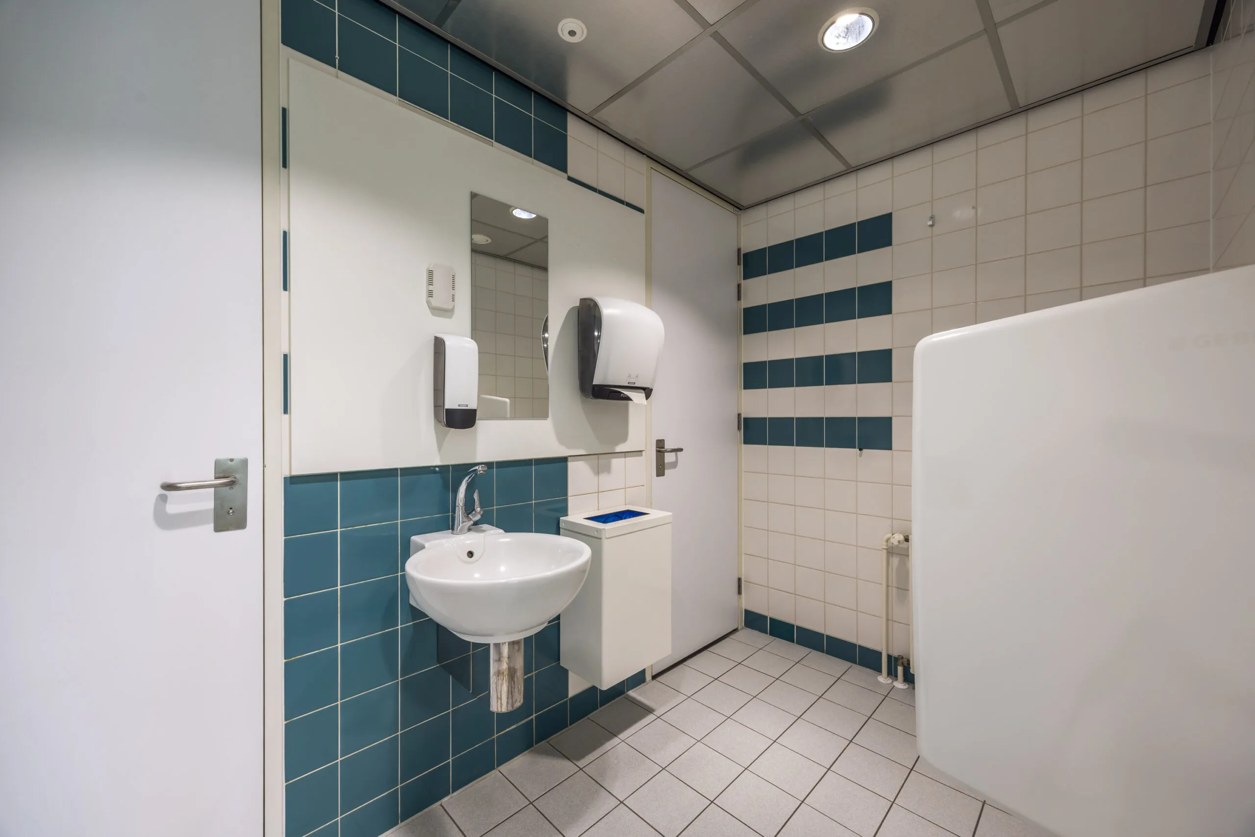 Moderne, betegelde toiletruimte op Daltonlaan met wastafel, spiegel en zeepdispensers.