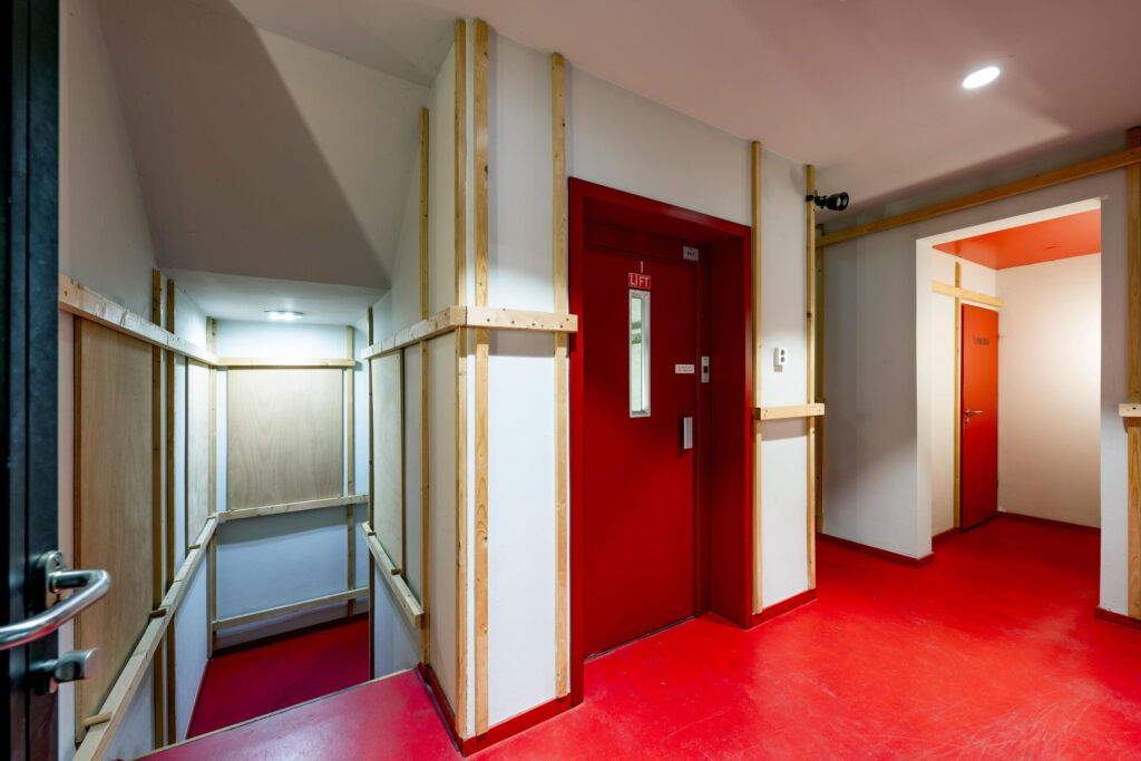 Hal met rode vloer en deuren, inclusief lift en trap, in een gebouw aan de De Ruijterkade.