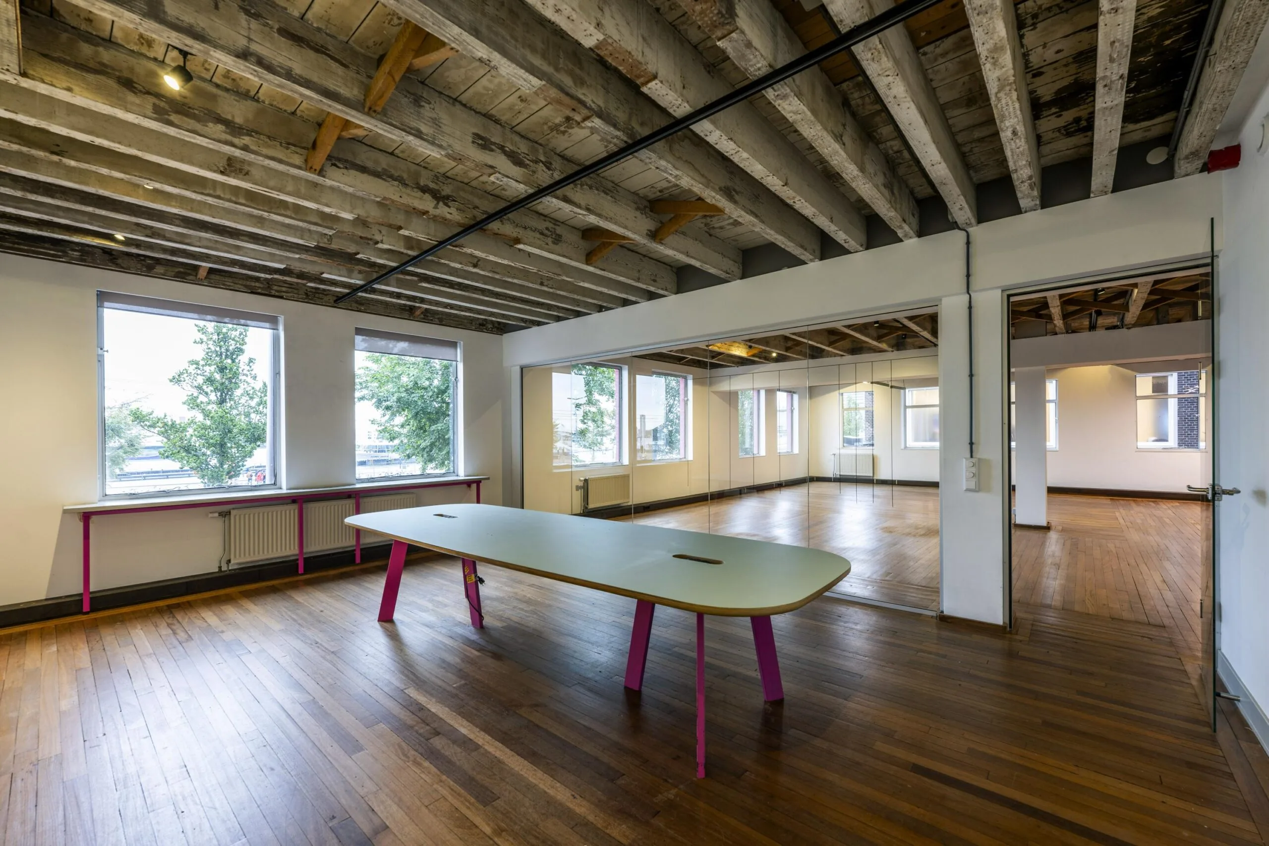 Modern kantoorinterieur aan de De Ruijterkade met houten vloer, zichtbare balkenplafonds en een vergadertafel met felroze poten.