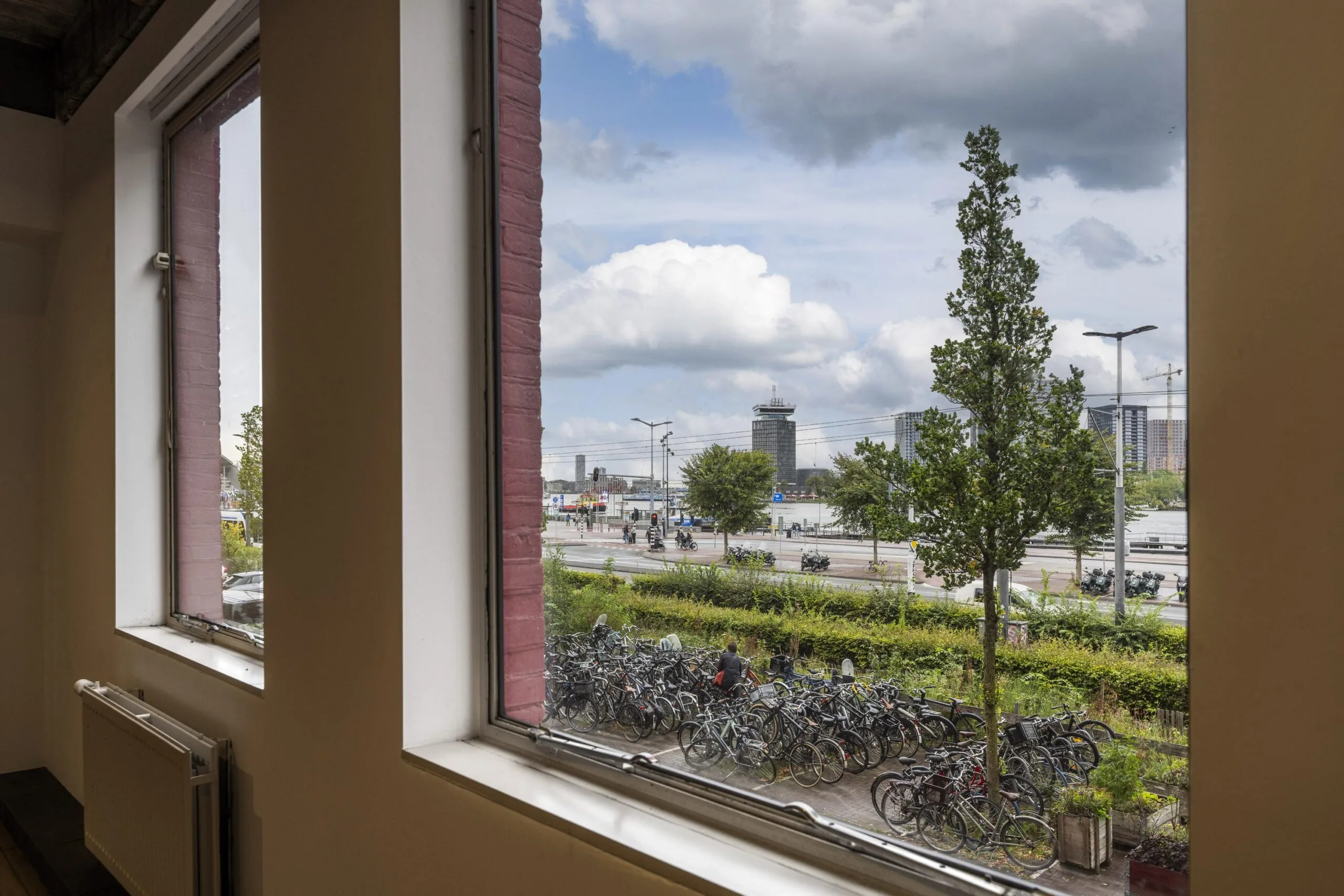 Uitzicht vanuit een raam aan de De Ruijterkade op fietsenstalling, wegverkeer en gebouwen aan het IJ in Amsterdam.