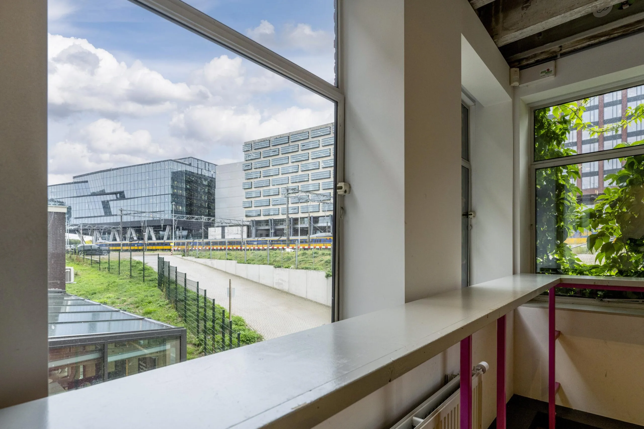 Uitzicht vanuit een binnenruimte op de moderne kantoorgebouwen aan de De Ruijterkade in Amsterdam, met een passerende trein op de voorgrond.