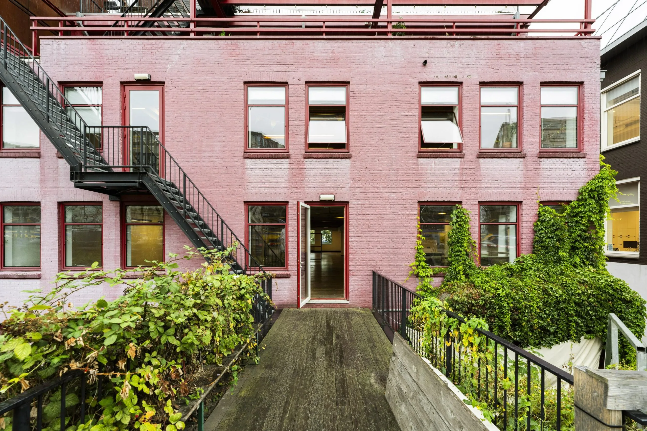 Vooraanzicht van een roze bakstenen kantoorgebouw aan de De Ruijterkade met rode kozijnen, buitentrap en begroeide gevel.