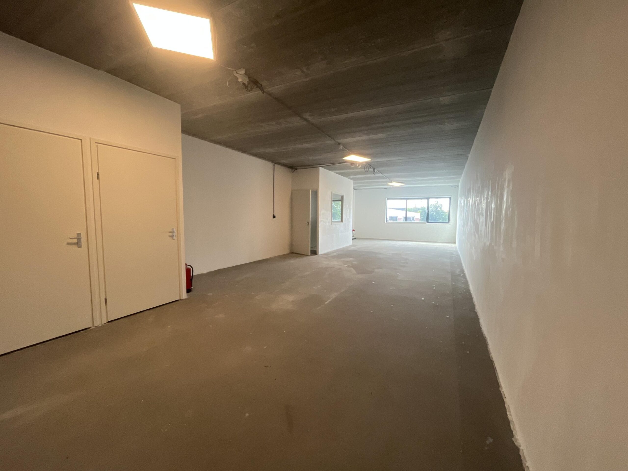 Een lege, onafgewerkte kamer met betonnen vloer en twee deuren aan de linkerkant.